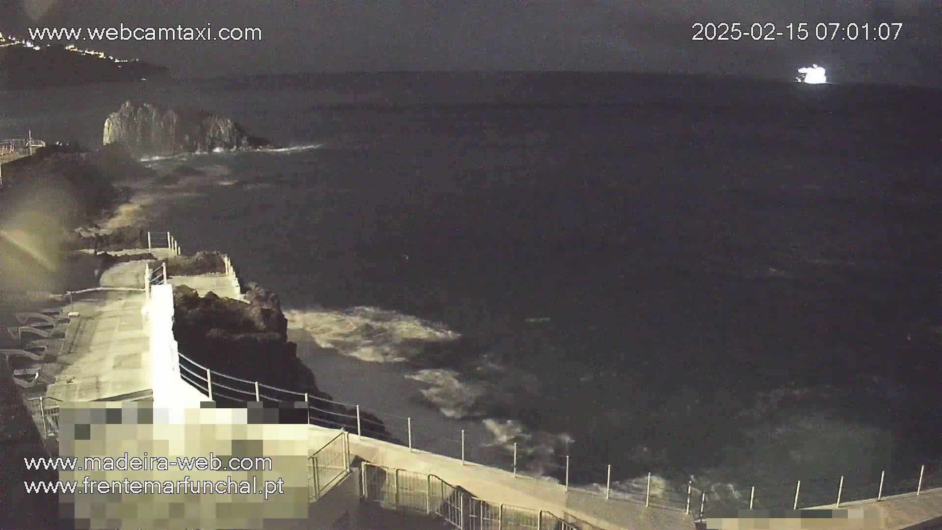 A nighttime seaside view shows dark, wavy water lapping against a concrete walkway and rocky shoreline.