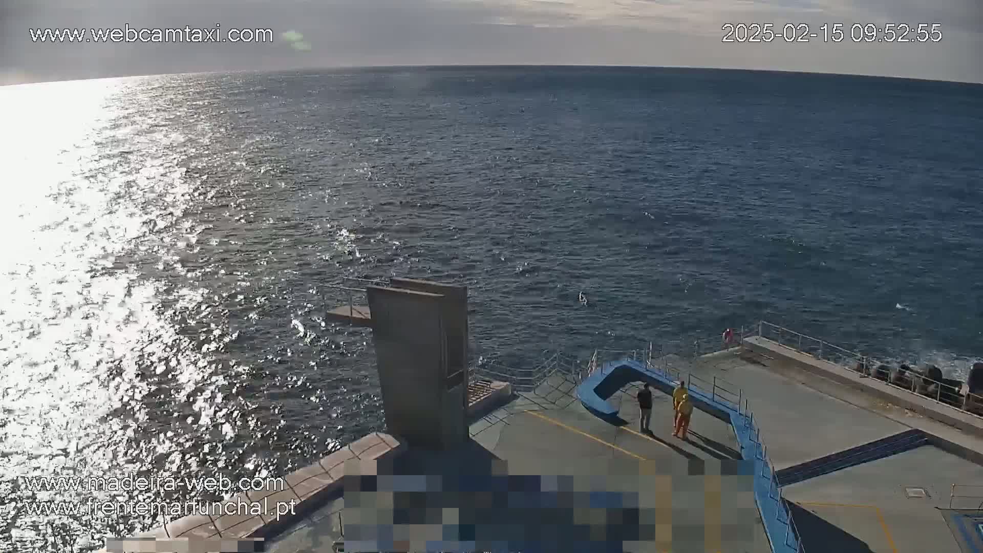 A high-angle view shows a partly sunny ocean with a diving board and a few people standing near a pool area on a concrete structure.