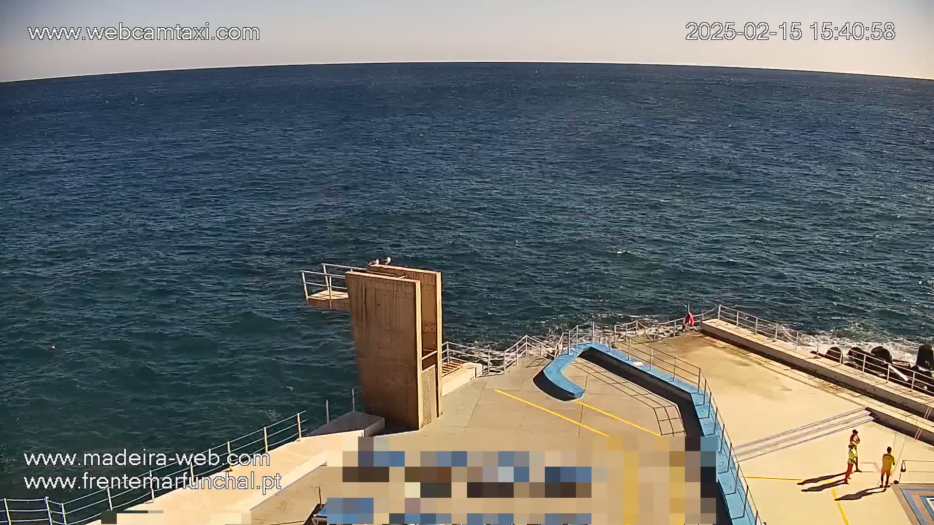 A high-angle, long shot on a sunny day shows a wooden diving platform overlooking a dark blue ocean with small waves lapping against a concrete seaside structure where a few people stand.