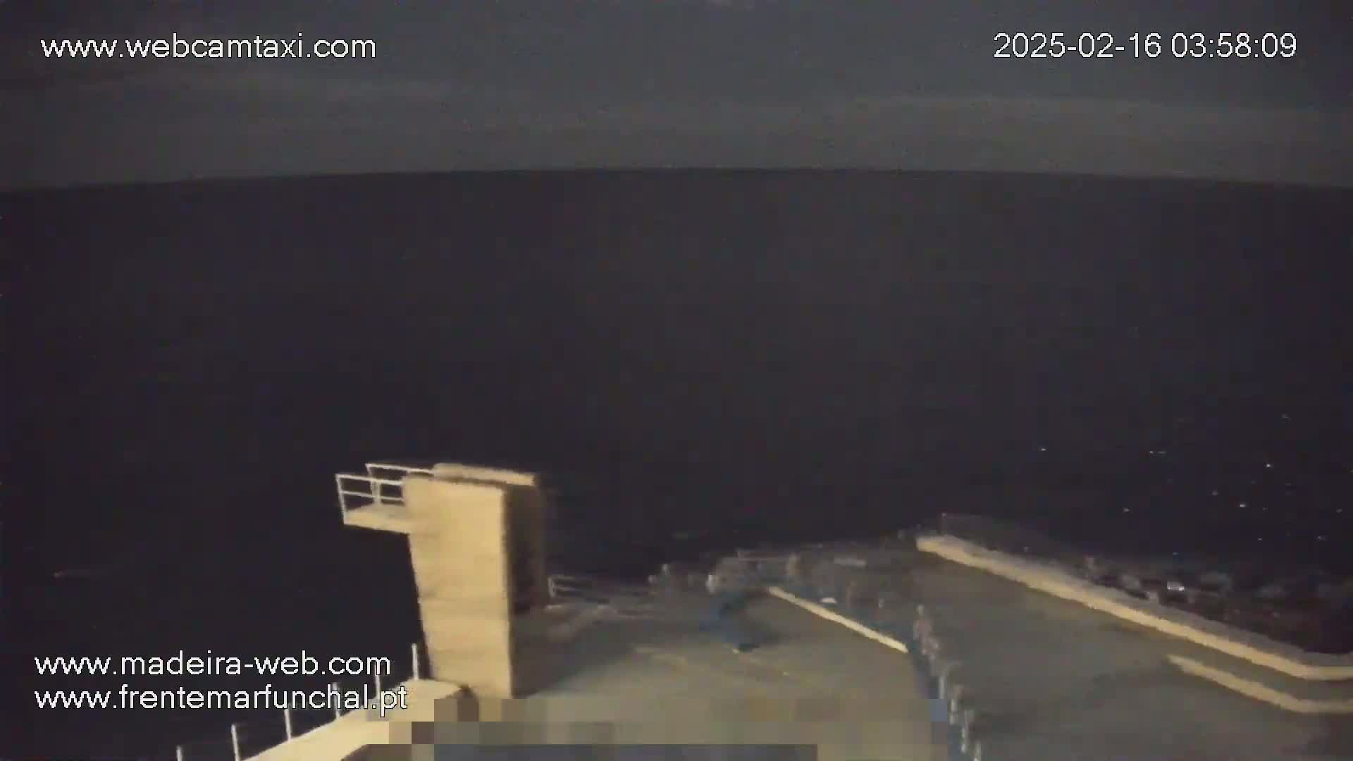 A nighttime view of a dark ocean from a high vantage point showing a diving board and several people on a concrete platform.