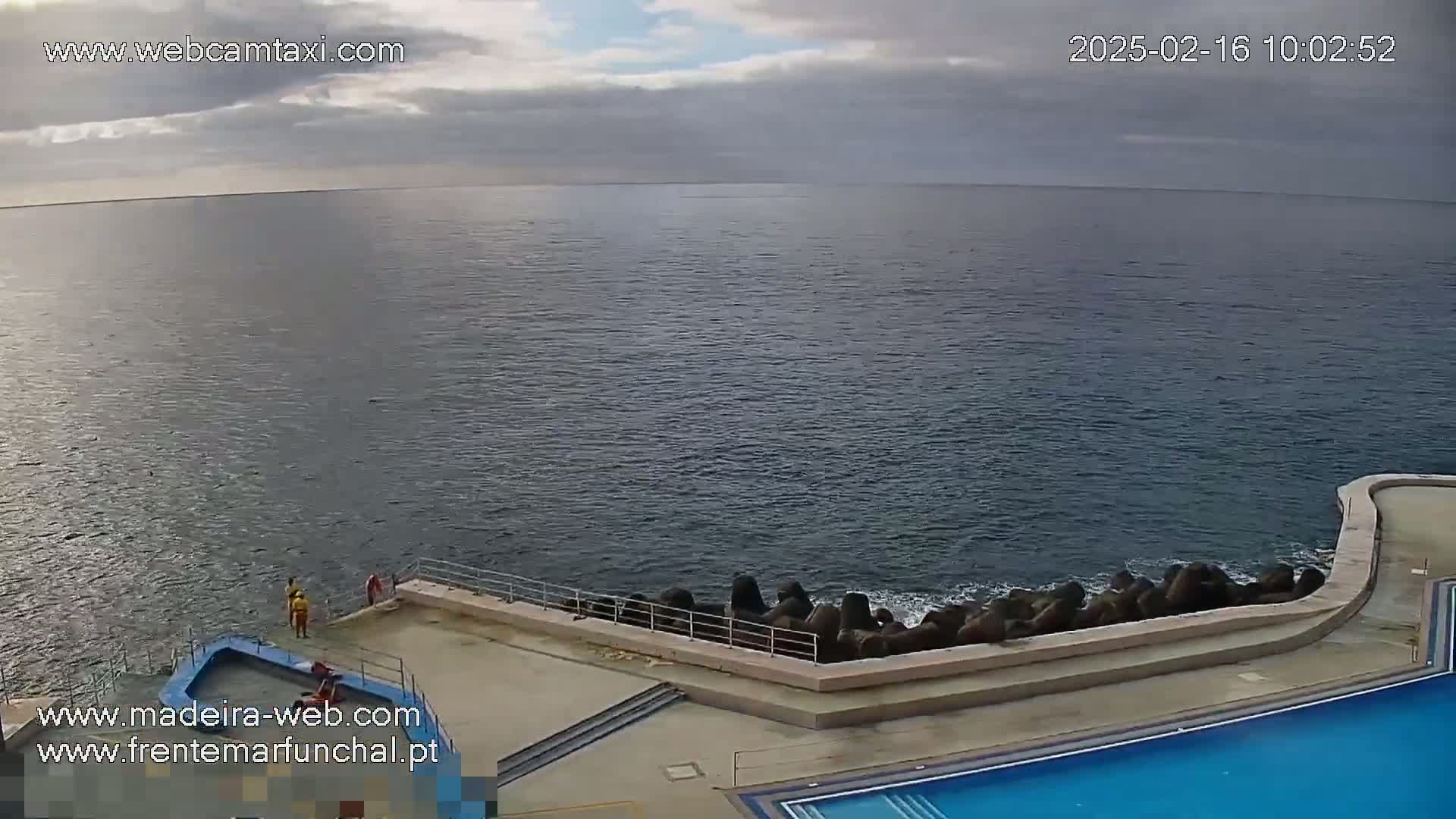 A mostly cloudy day shows a calm ocean meeting a concrete seawall and pool area, where a few people are standing near a small boat.