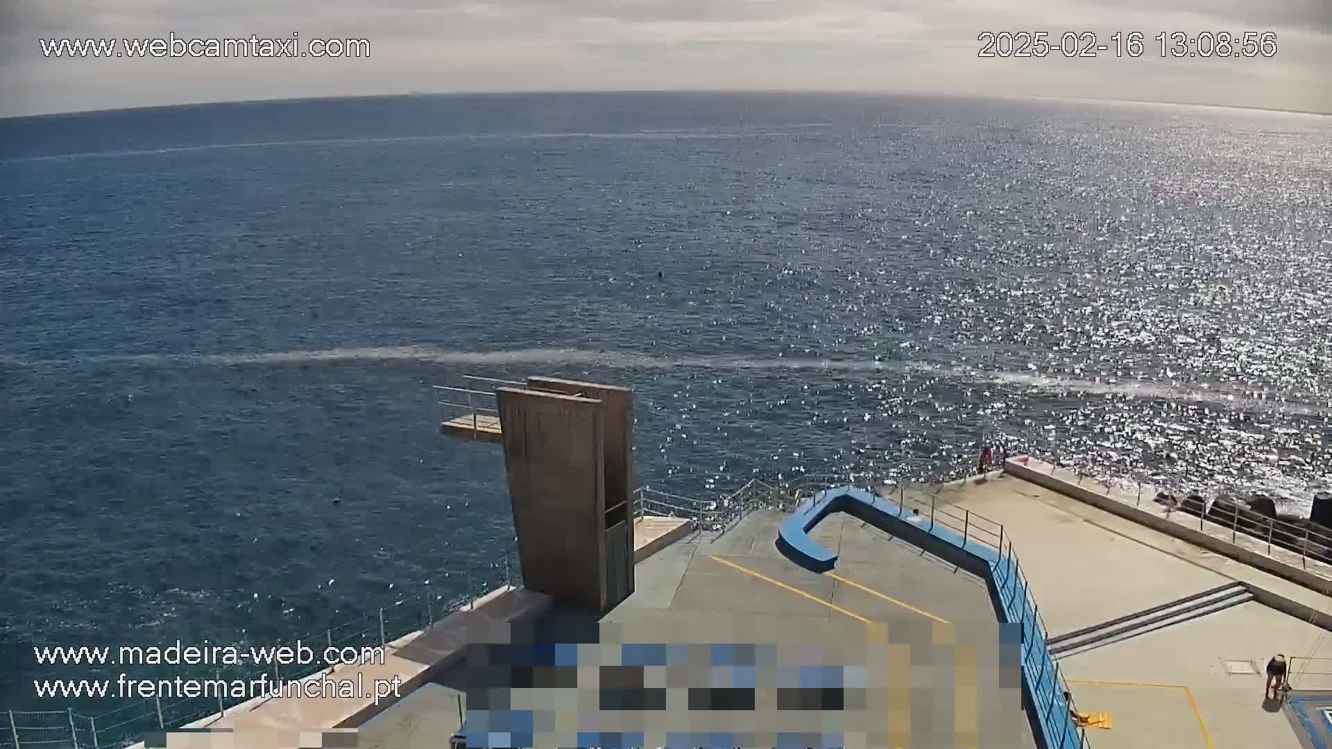 A high-angle view shows a diving platform overlooking a sunlit, wavy ocean on a partly cloudy day.