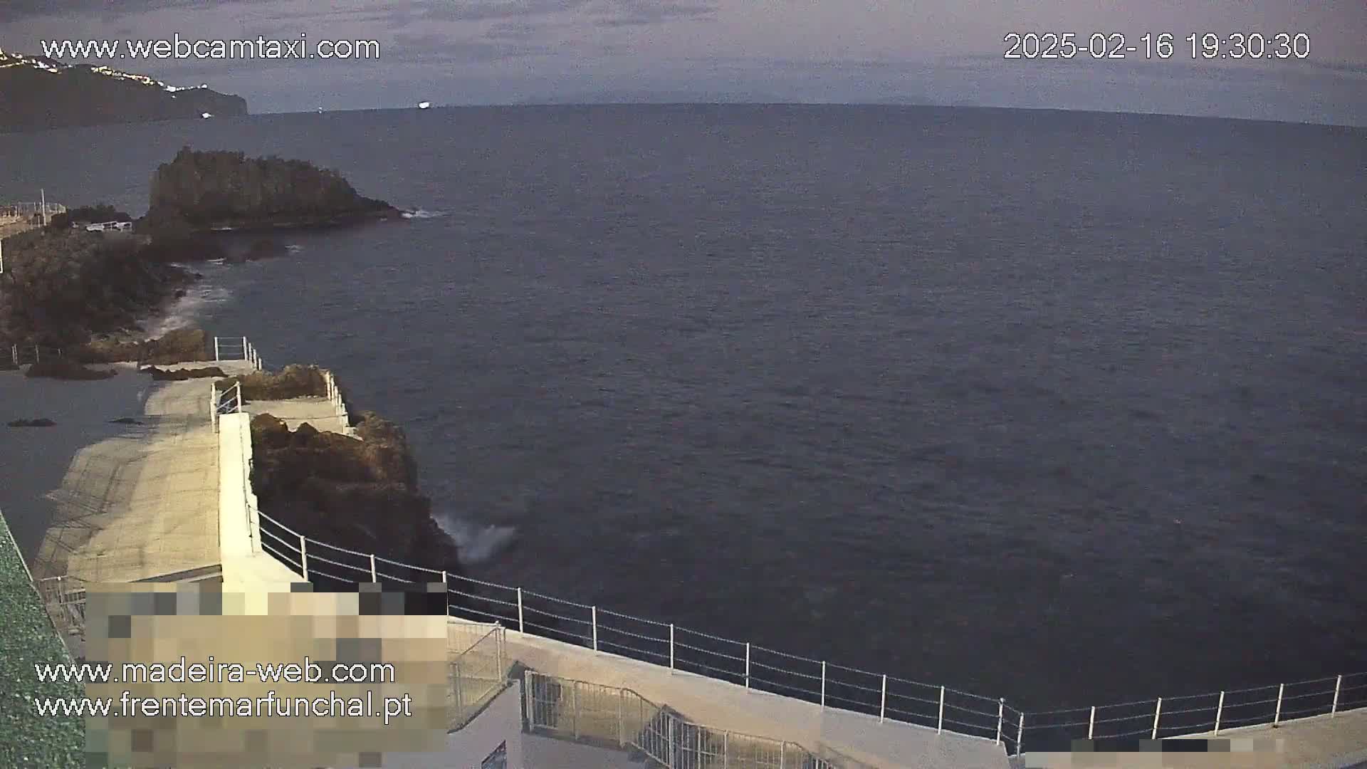 A concrete walkway along a rocky coastline overlooks a dark, choppy ocean under a cloudy sky.