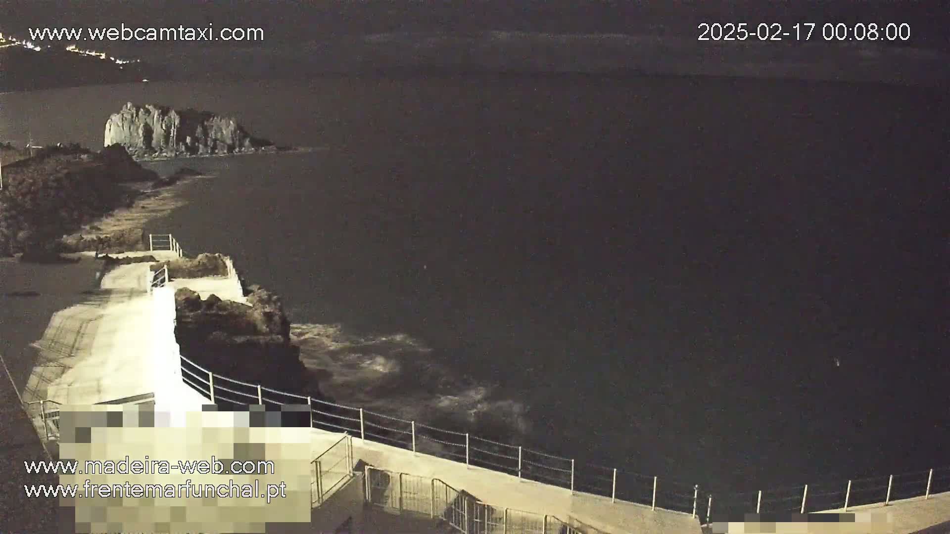 A nighttime coastal scene shows a rocky outcropping in dark water with gentle waves lapping against a concrete walkway and railing.