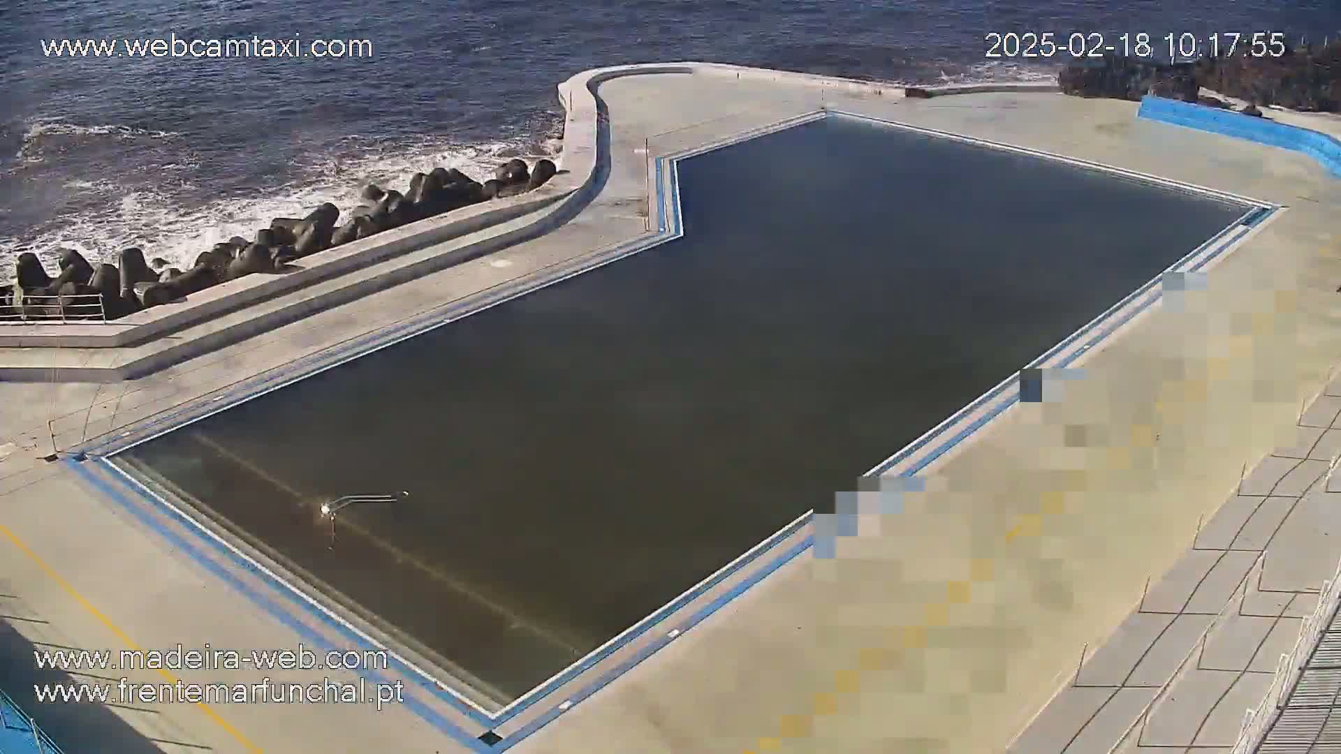 An outdoor oceanside swimming pool with dark water is surrounded by concrete and a rock barrier against the ocean waves.
