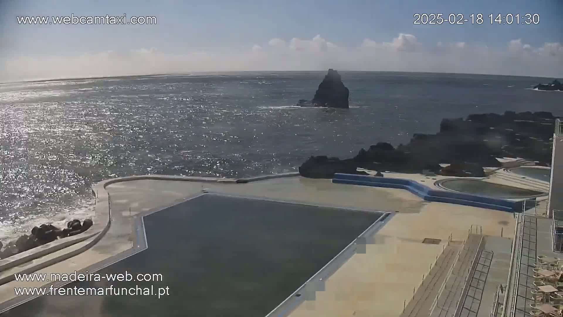An outdoor, oceanside pool complex with dark water, partially visible rock formations in the sea, and a mostly sunny sky.