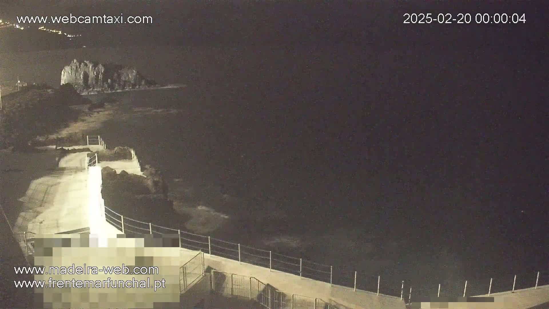 A nighttime coastal scene shows a paved walkway with a railing overlooking dark water and a rocky outcrop in the distance under a dark sky.