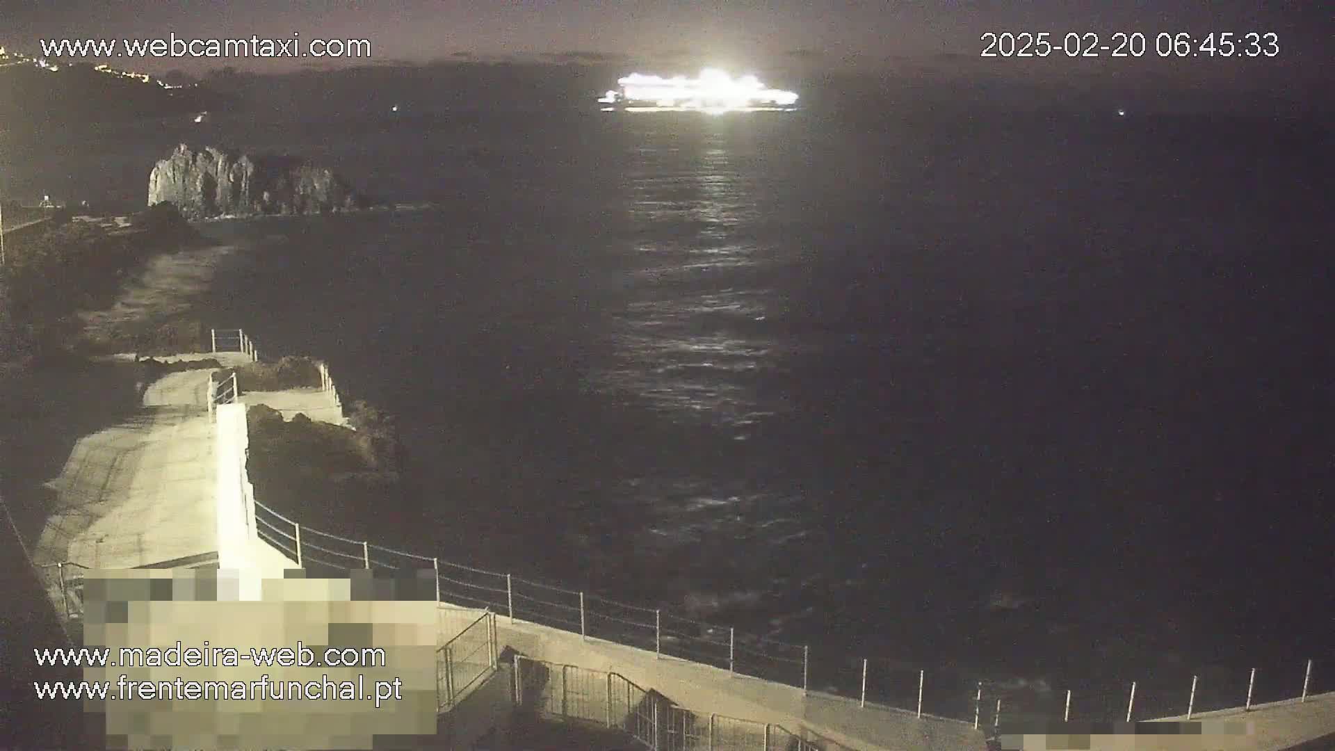 At night, a brightly lit ship is visible on dark water from a high vantage point overlooking a paved walkway and a rocky shoreline.