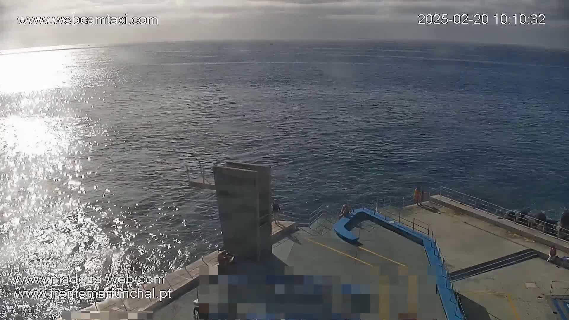 A mostly sunny day shows a rocky coast with a diving platform and several people standing near a blue railing overlooking a dark blue ocean with some wave spray.