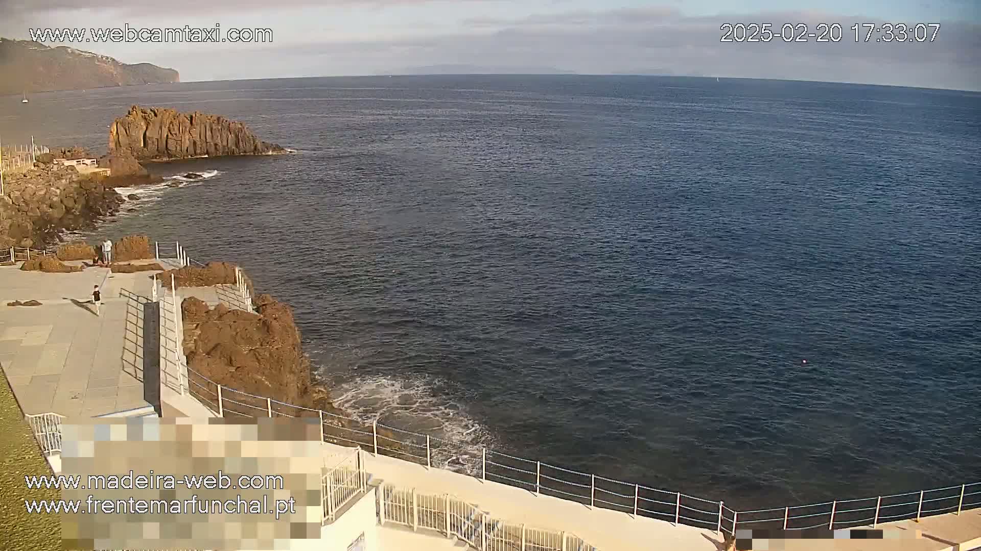 A rocky coastline with a paved walkway and railing overlooks a calm, dark blue ocean under a partly cloudy sky.