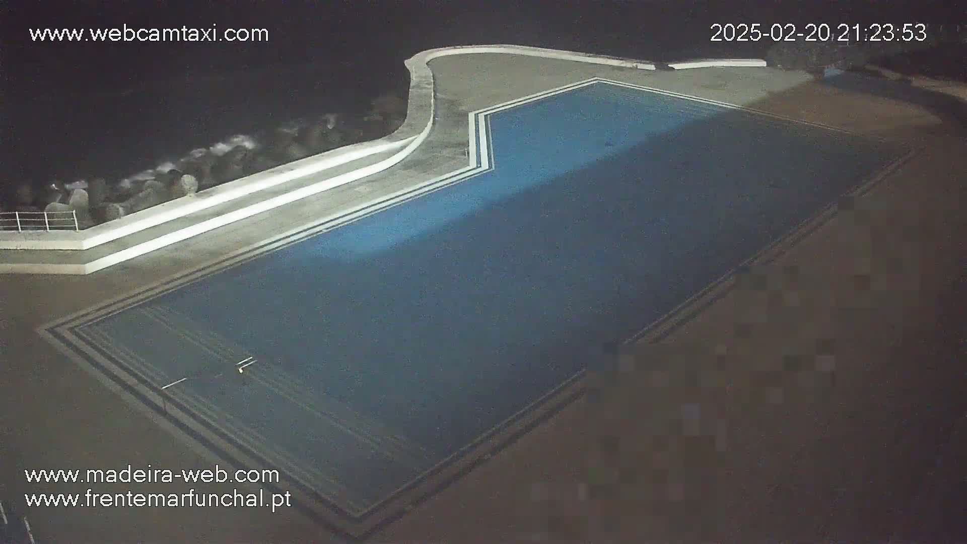 An outdoor nighttime view of a dark blue rectangular swimming pool next to a rocky ocean.