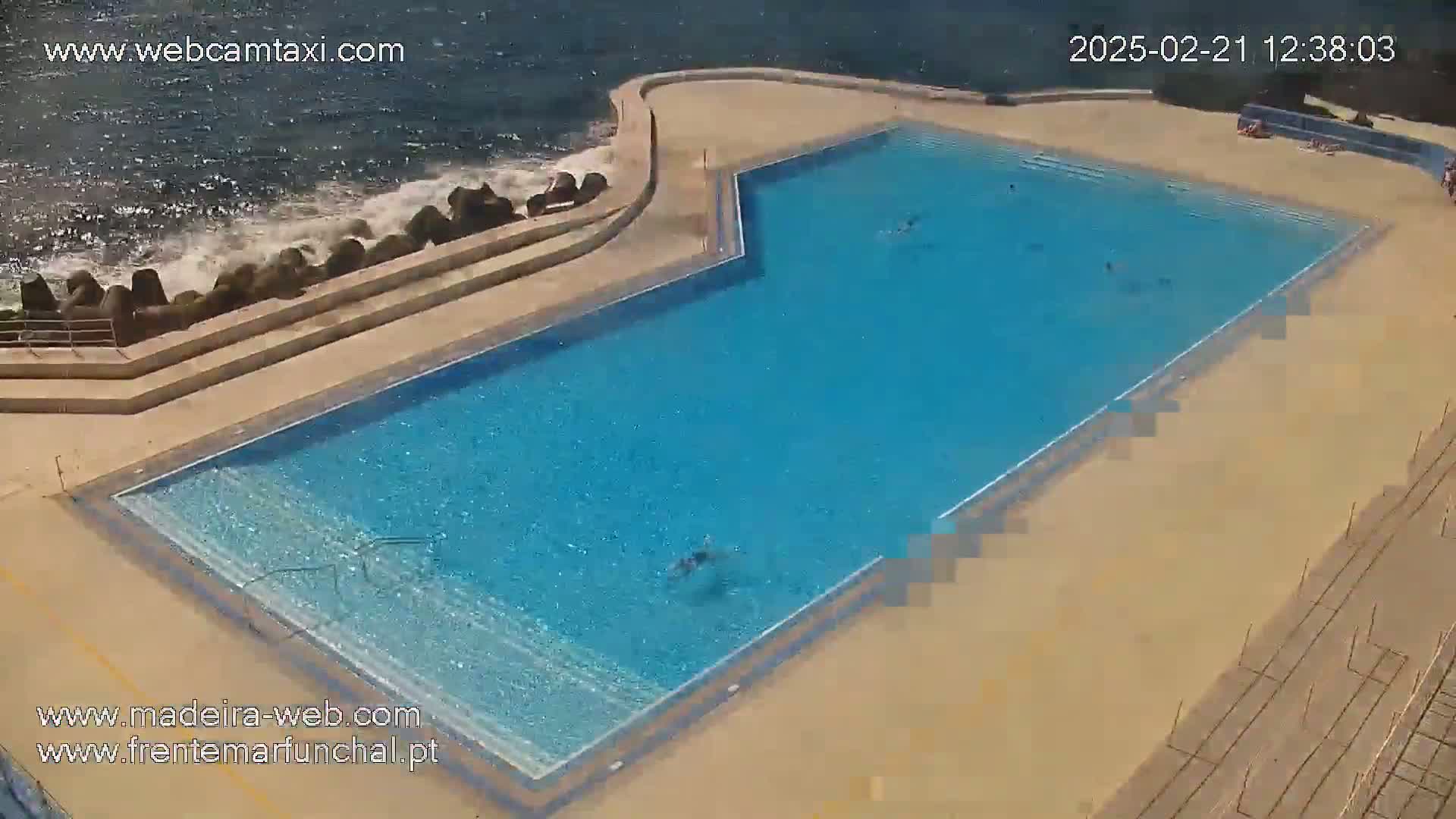 An outdoor swimming pool with several people swimming in it is situated next to a rocky ocean shore on a sunny day.