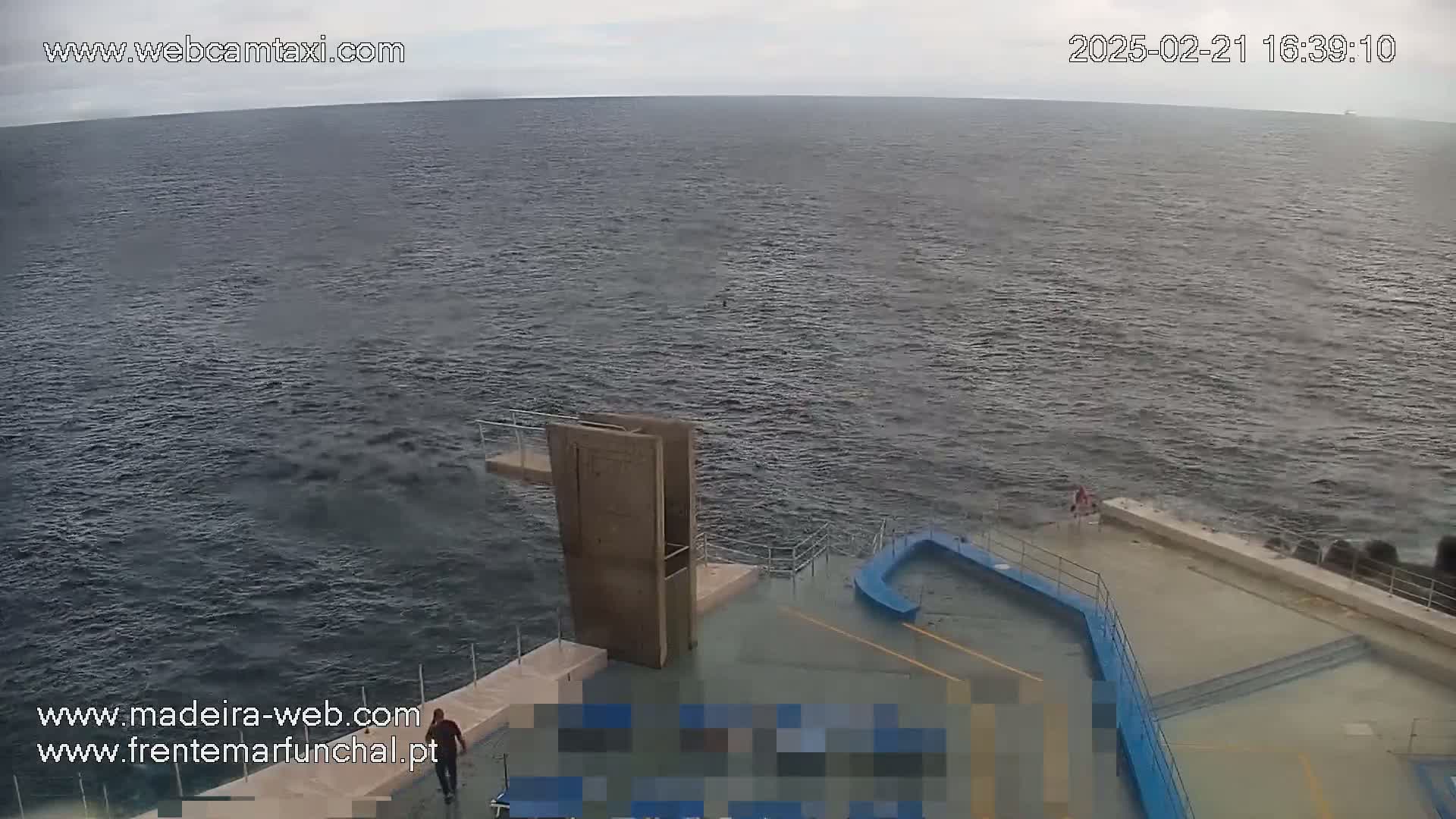 A person stands near a diving platform overlooking a dark, choppy ocean under an overcast sky.