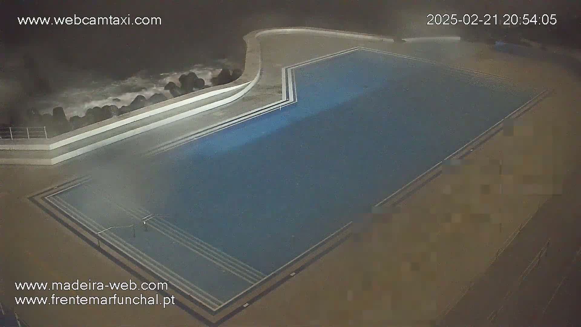 An outdoor nighttime aerial view shows a dark-blue swimming pool next to a rocky coast with crashing waves.