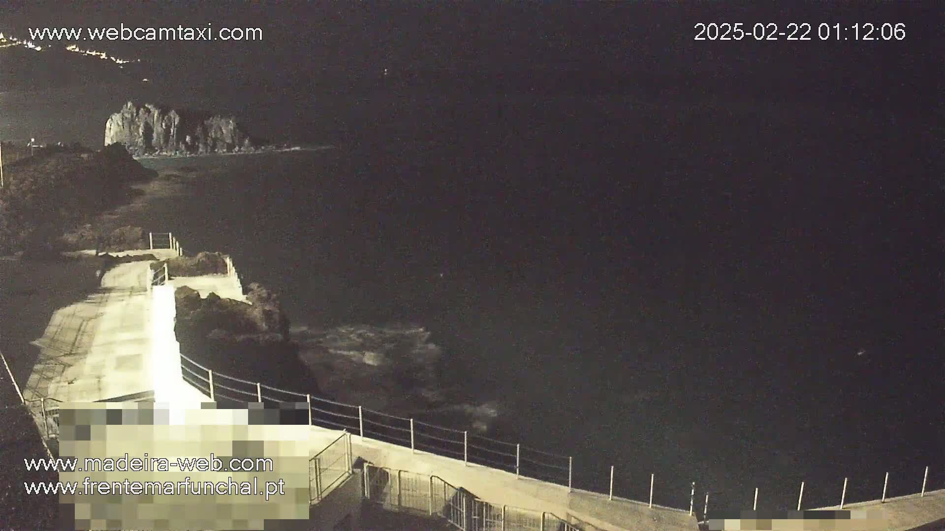 A nighttime, coastal scene shows a paved walkway with a railing overlooking dark ocean water and rocky formations.