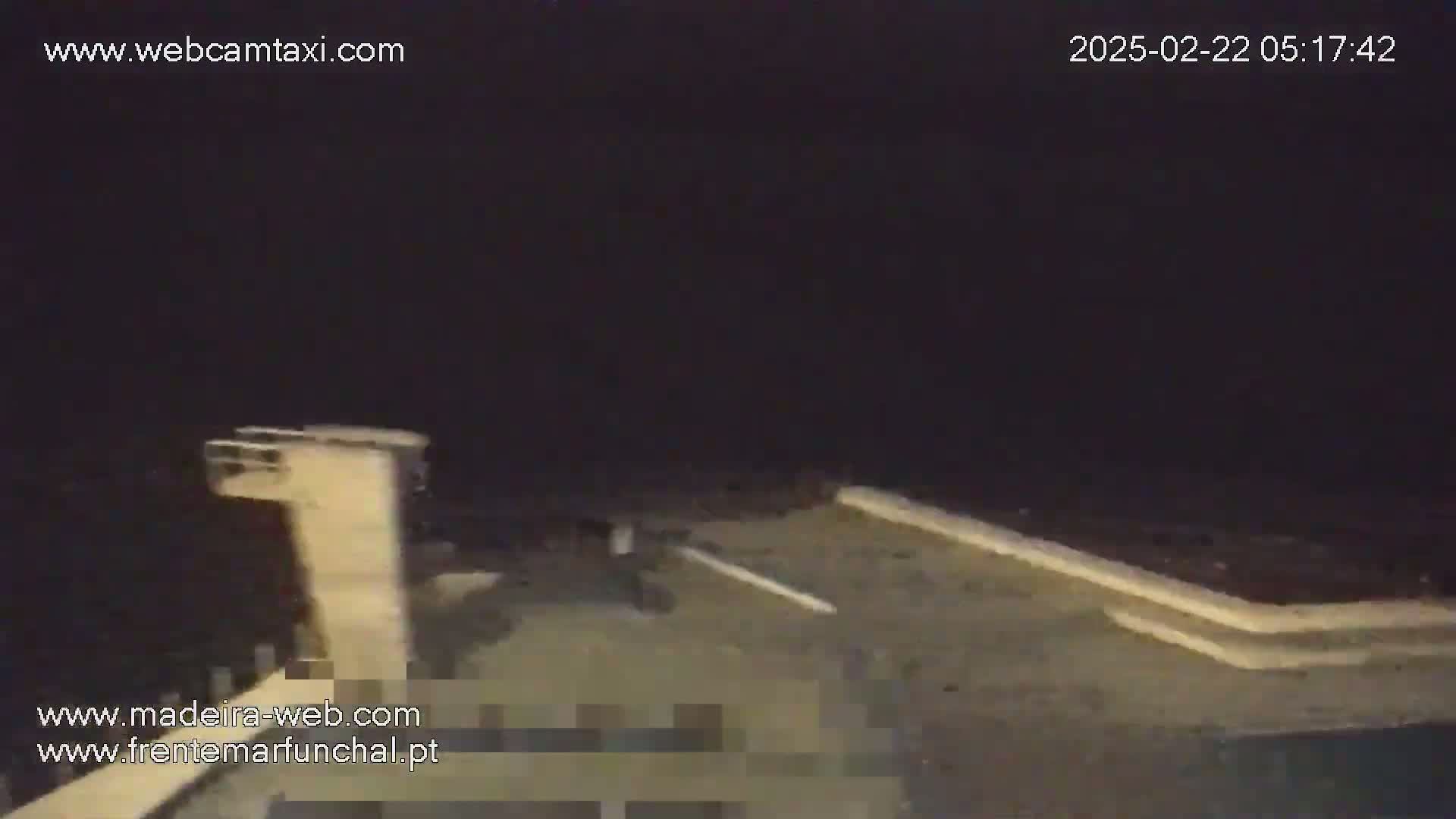 A nighttime, outdoor scene shows a section of a light-colored wall and ground, possibly near a body of water, under a dark sky.