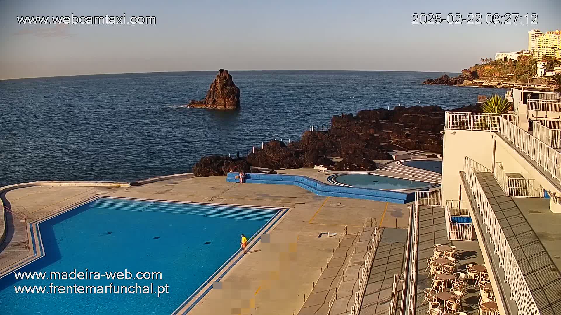 An outdoor pool complex with a large blue swimming pool and smaller circular pool overlooks a dark-colored rocky coastline and ocean under a clear, bright sky.