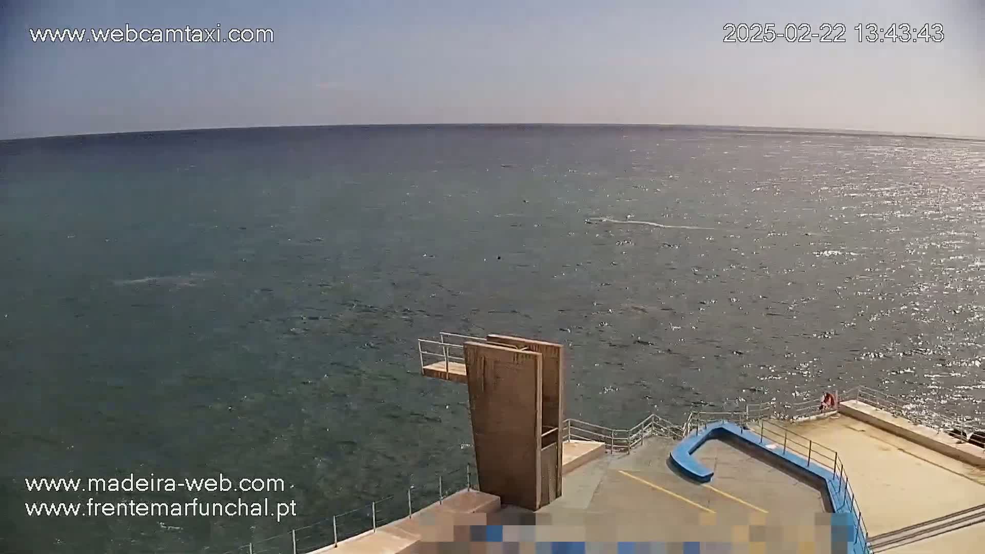 A high-angle view shows a diving platform overlooking a calm, slightly rippled ocean under a clear sky.
