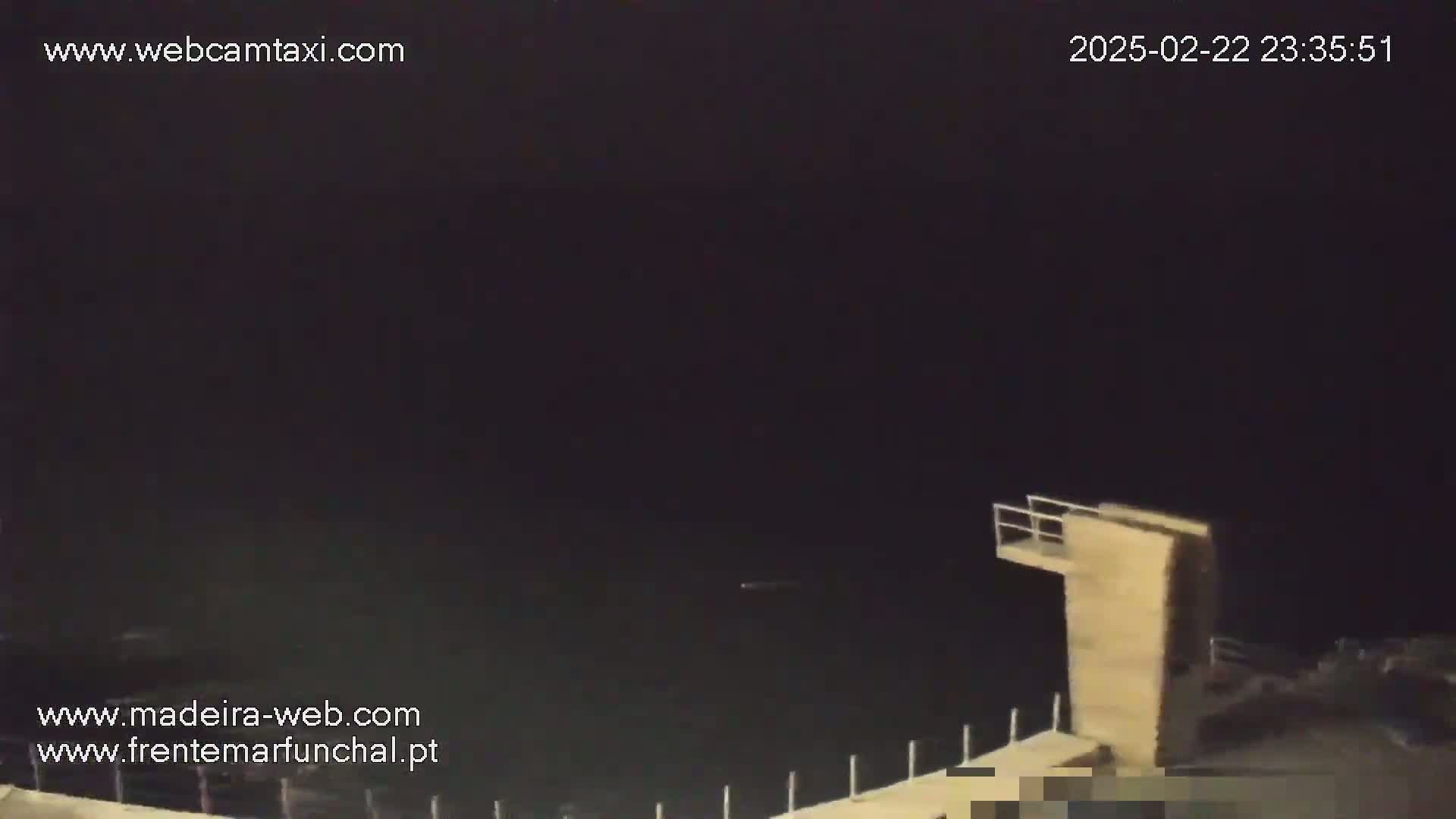 A nighttime view of dark water, a diving platform, and a low-lit shoreline under a dark sky.