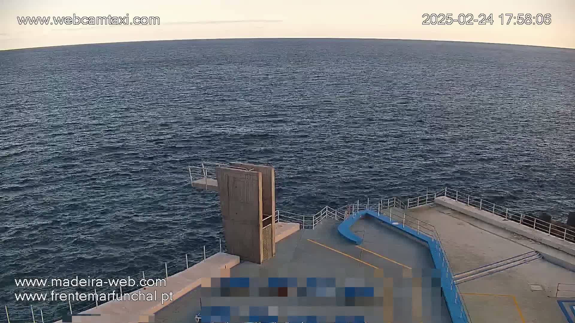 A high-angle, long shot shows a diving platform overlooking a calm, dark-blue ocean under a pale, clear sky at dusk.