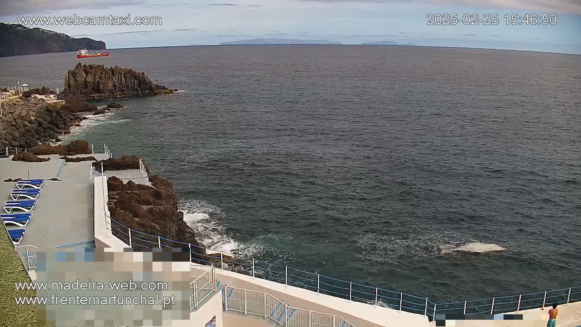 A mostly cloudy day shows a rocky coastline with a sunbathing area overlooking a dark-blue ocean where a small vessel is visible in the distance.