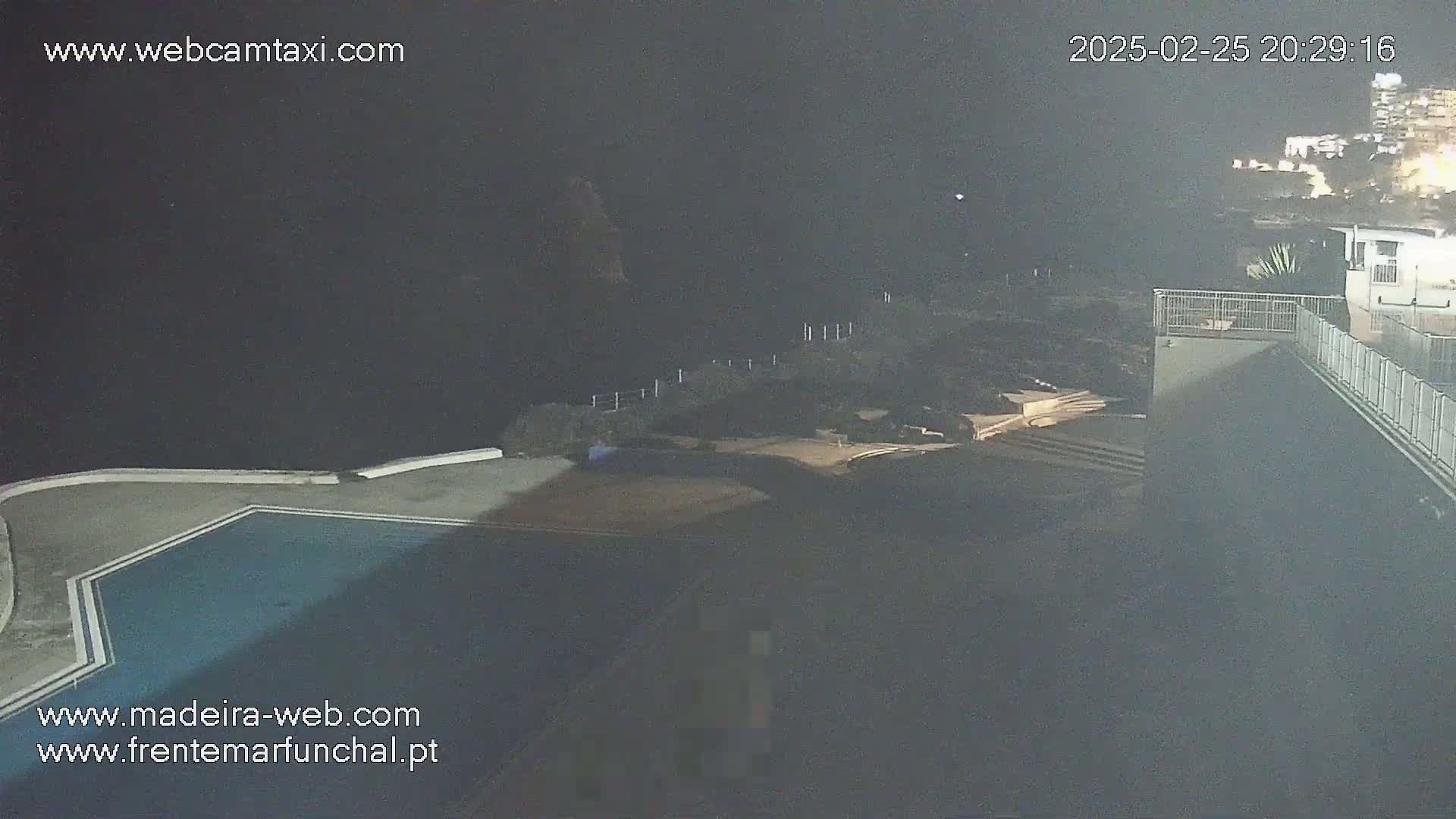 A nighttime, high-angle view shows an empty swimming pool and surrounding structures next to a dark ocean.