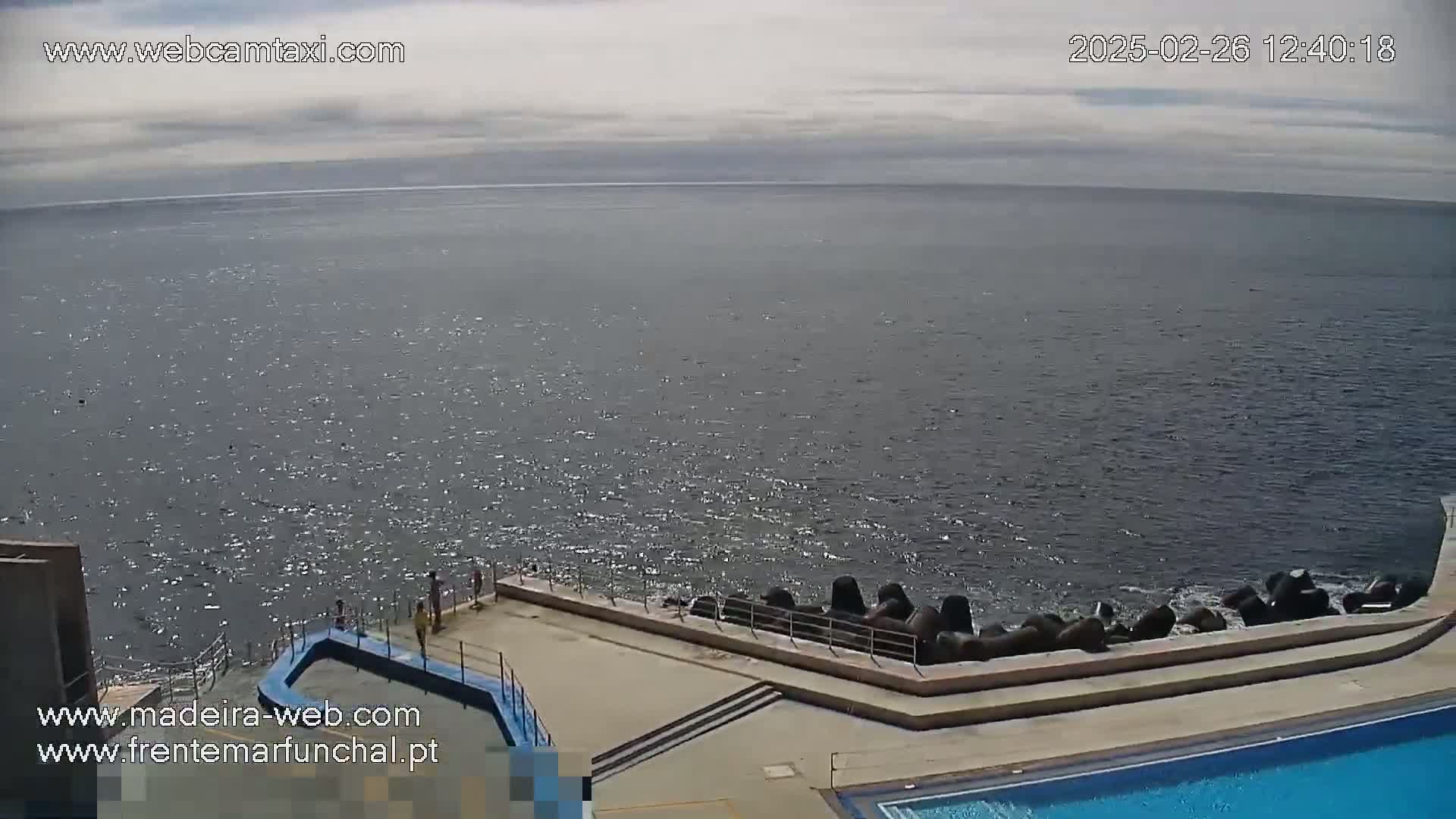 A mostly cloudy sky overlooks a calm ocean meeting a concrete shoreline with a pool and a few people near the water.