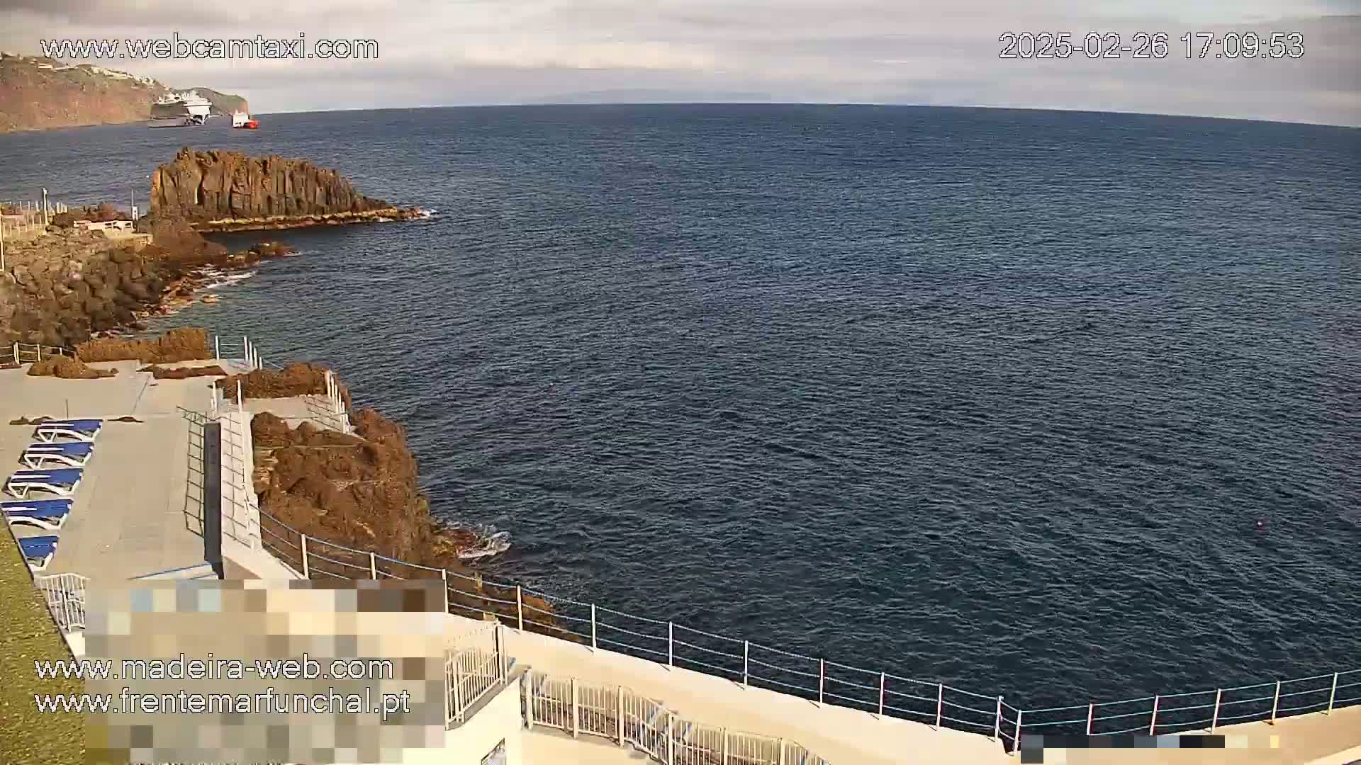 A mostly sunny day overlooking a dark blue ocean with rocky coastlines and lounge chairs on a deck, and a large ship visible in the distance.