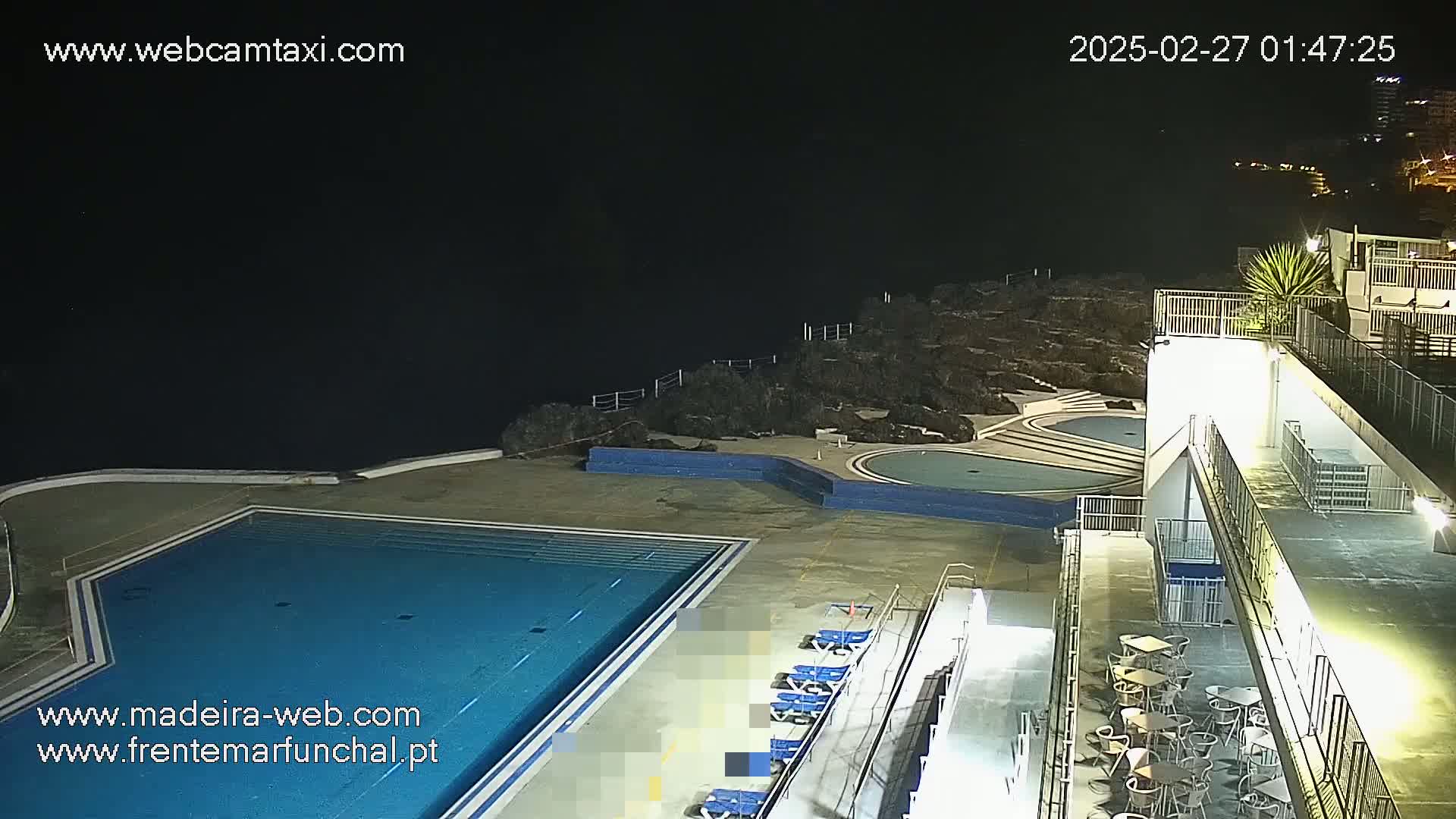An outdoor nighttime scene shows an empty swimming pool complex with lounge chairs, some tables and chairs, and a rocky cliff overlooking a dark ocean.