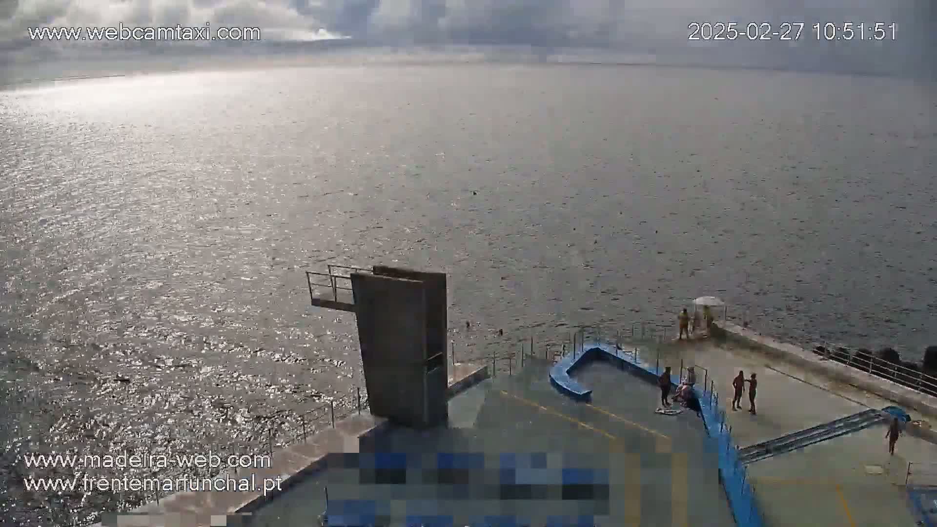 A mostly sunny day overlooking a calm ocean with a diving platform and several people on a concrete deck.