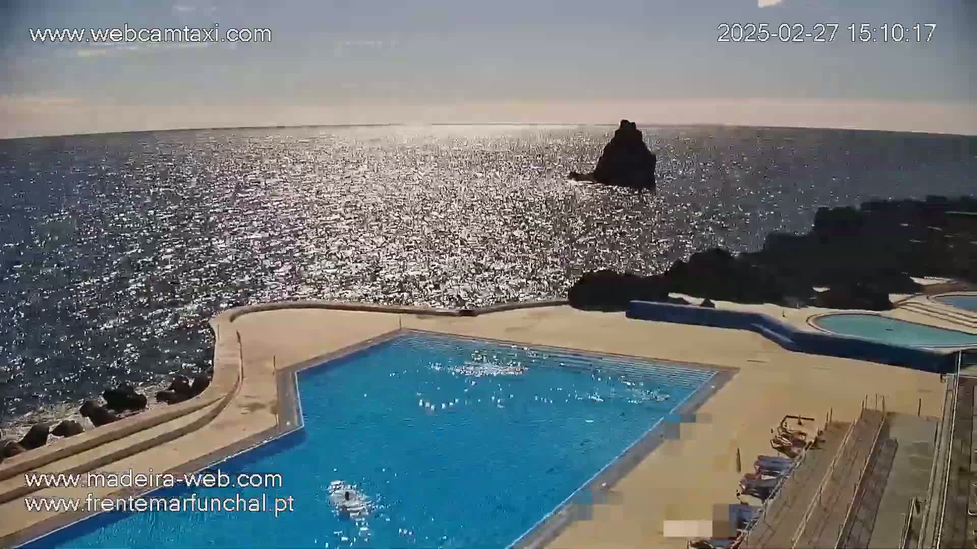 A large, rectangular swimming pool is situated on a cliff overlooking a sparkling ocean under sunny skies.