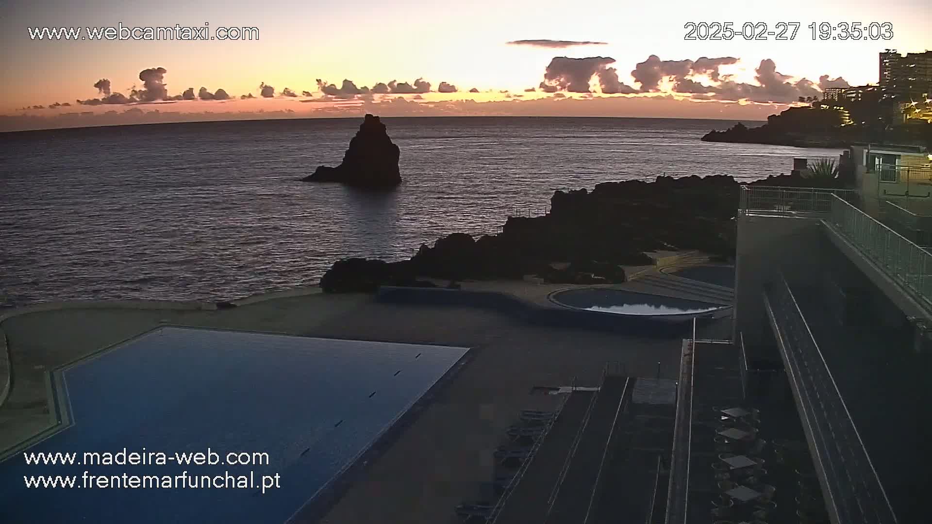 An ocean sunset illuminates a seaside resort with an infinity pool overlooking dark volcanic rock formations and calm water.