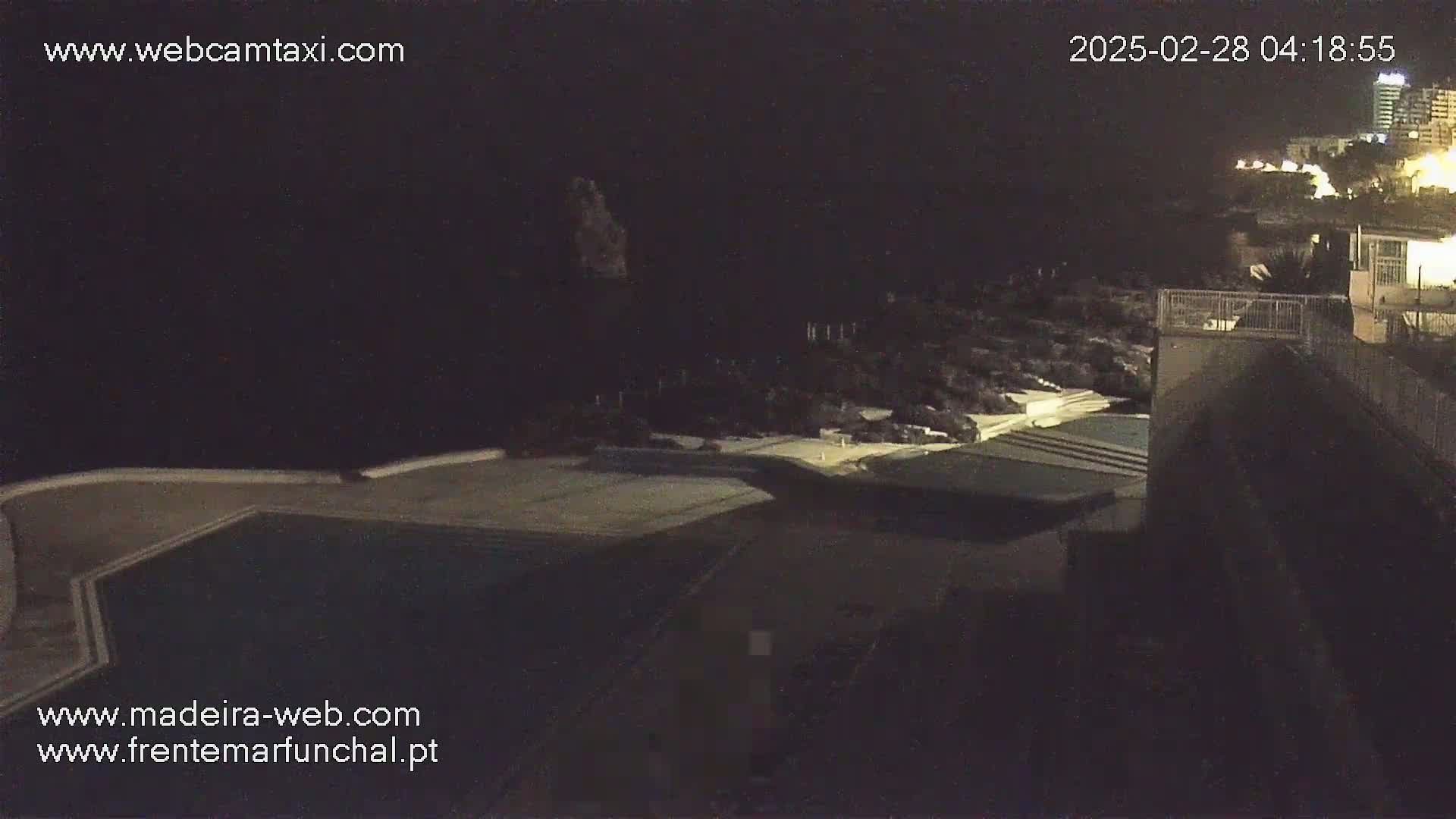 A nighttime, high-angle view shows an outdoor swimming pool complex situated near a dark ocean and a dimly lit coastline.