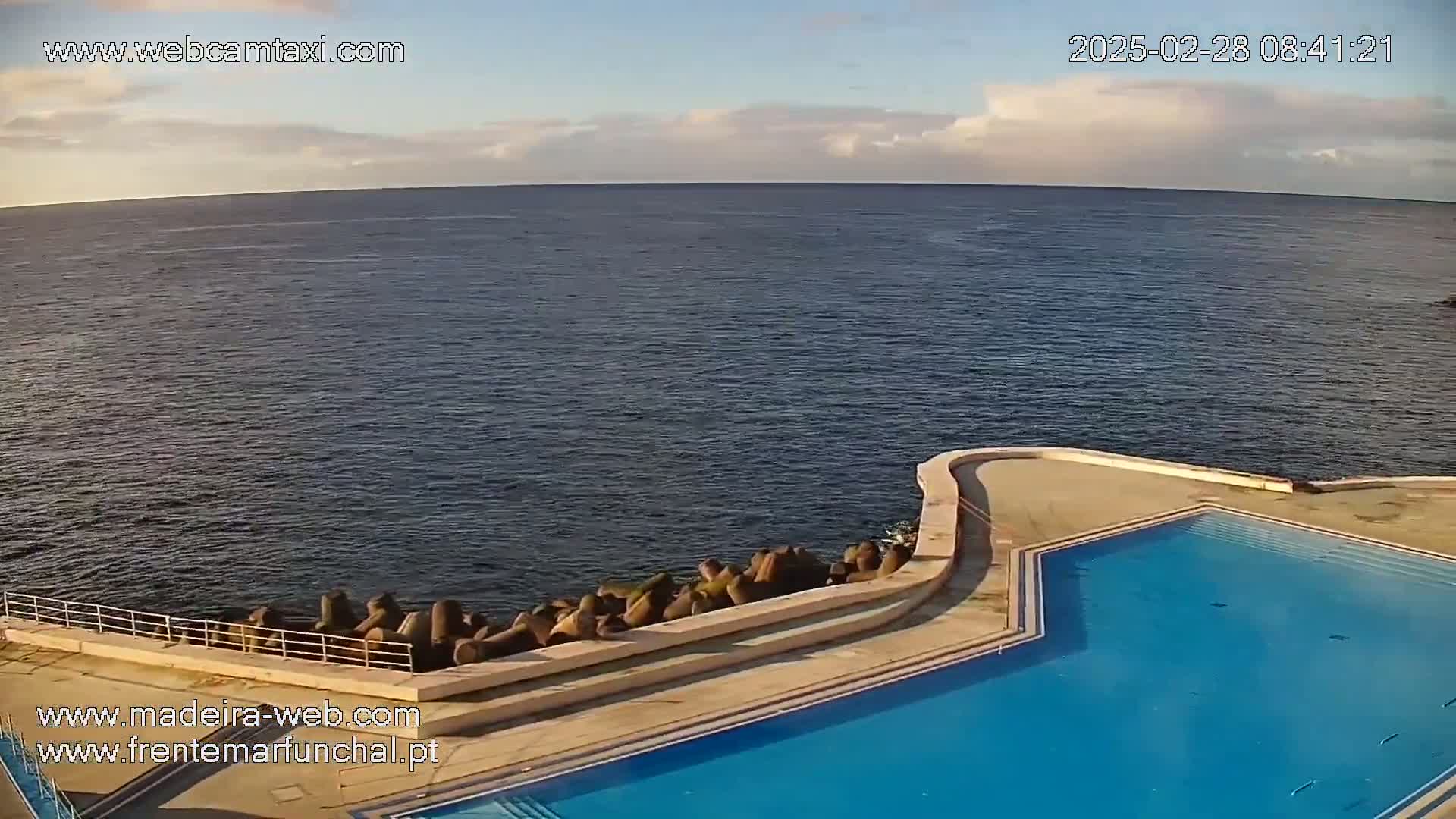 An outdoor swimming pool complex next to a calm ocean under a partly cloudy sky.