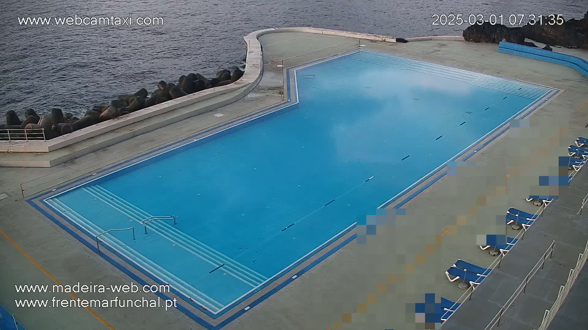 An outdoor, blue swimming pool with several empty lounge chairs sits next to a dark body of water.