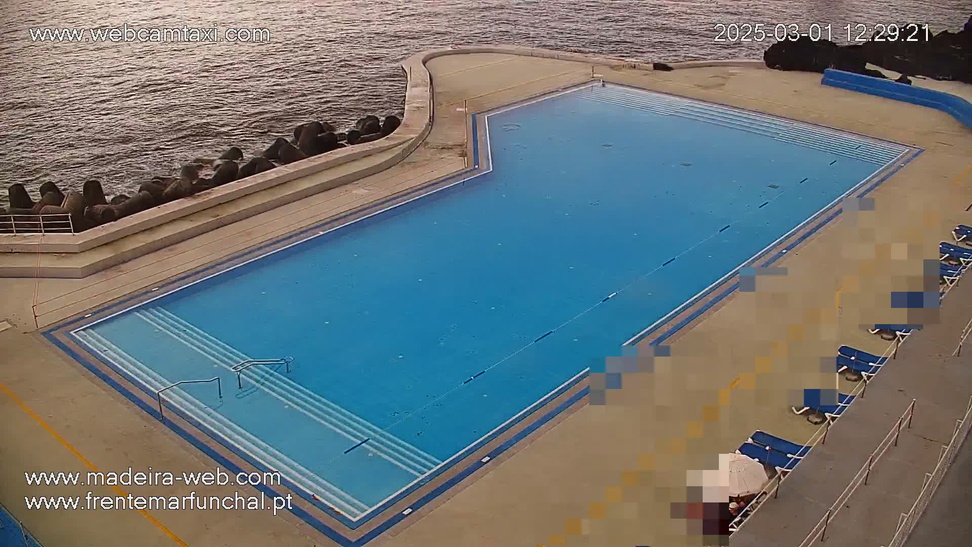 An outdoor pool with several lounge chairs sits next to a rocky ocean shore on a calm day.