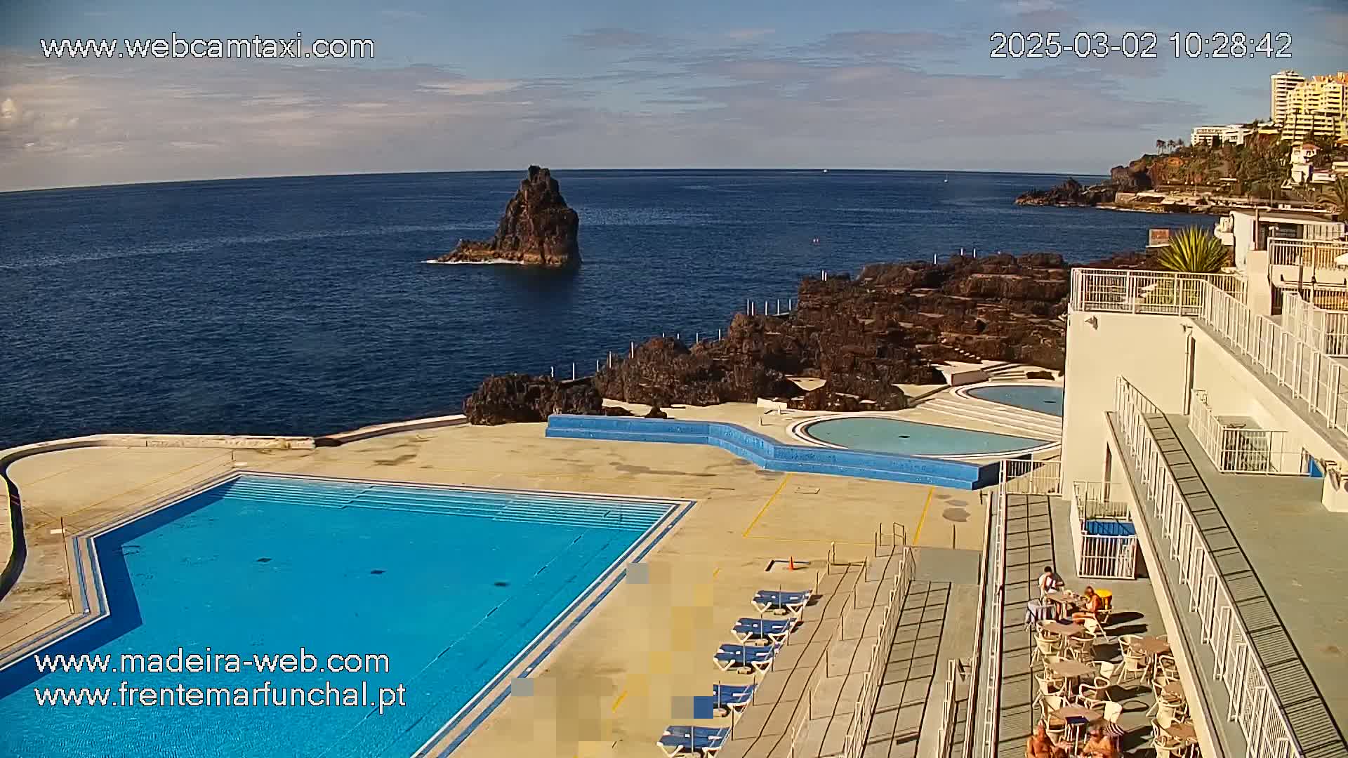 An outdoor pool complex sits on a rocky coast overlooking a calm ocean, under a mostly clear sky.