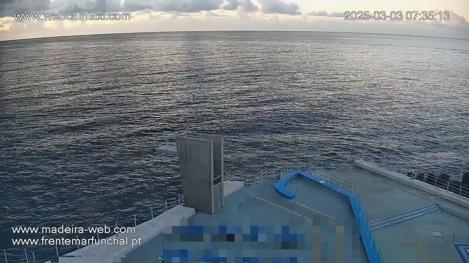 A mostly cloudy sky overlooks a calm, dark-blue ocean meeting a concrete structure with a diving platform.