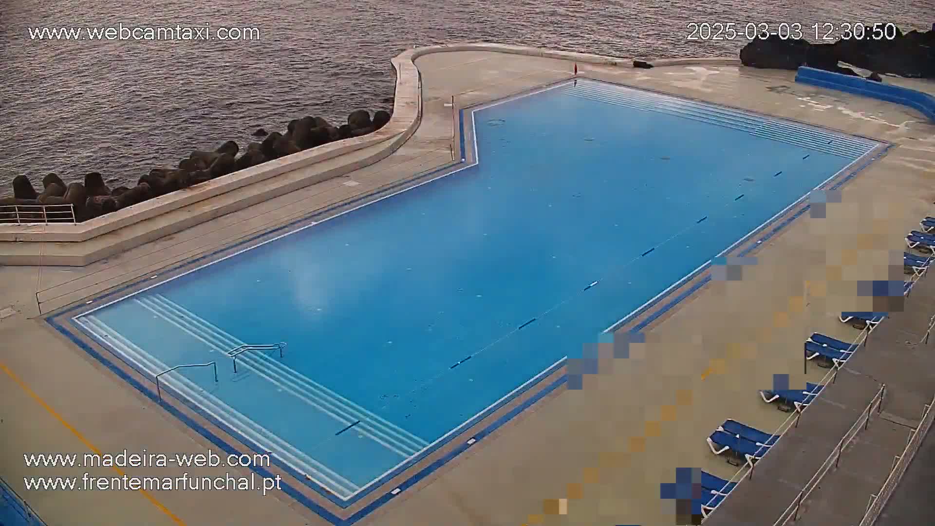 An outdoor, oceanside swimming pool with lounge chairs sits next to a rocky coastline under a mostly clear sky.