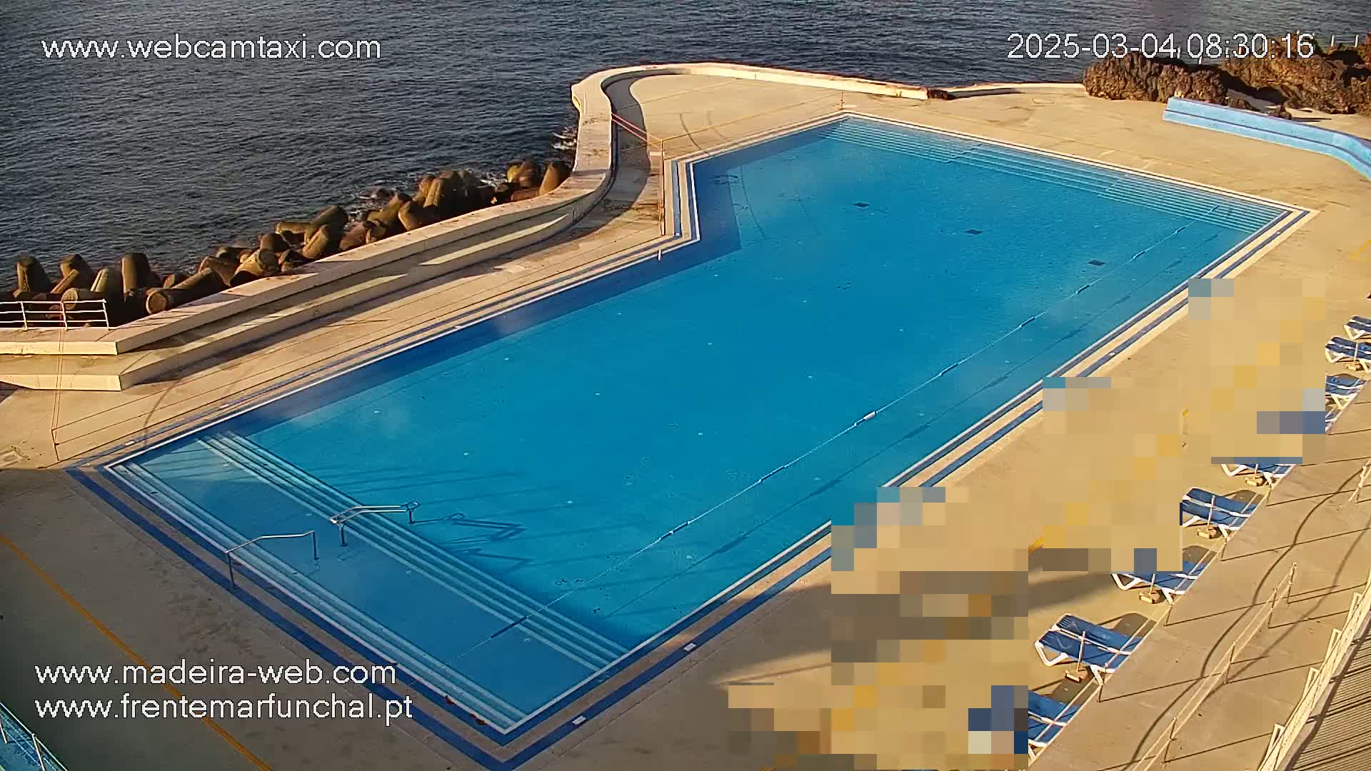 An outdoor, blue, rectangular swimming pool with several empty lounge chairs sits next to a calm ocean.
