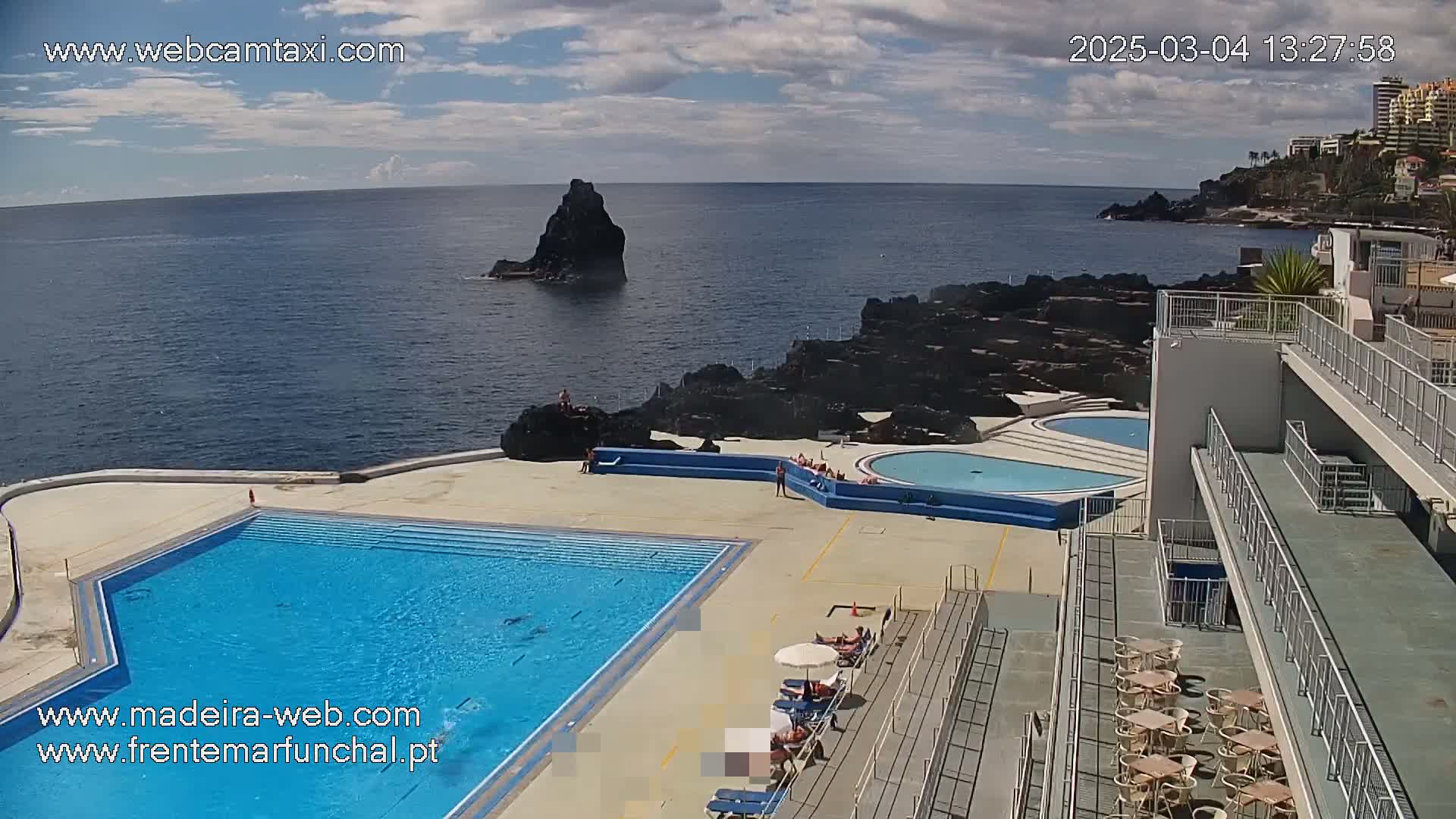 An outdoor pool complex with a large swimming pool and smaller pools overlooks a rocky coastline and ocean under a partly cloudy sky.