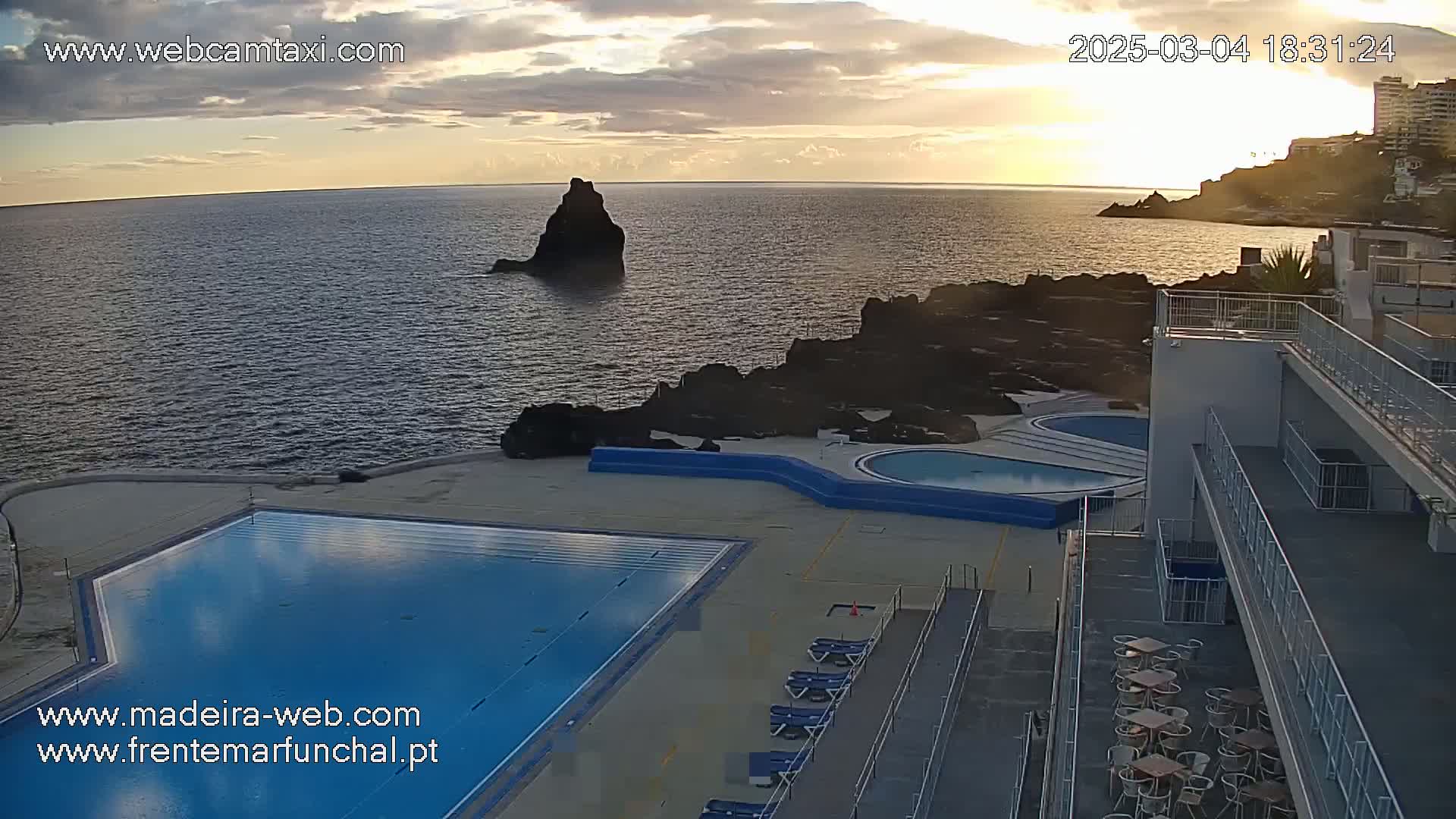 An ocean view at sunset shows a swimming pool and patio area next to a multi-story building with outdoor seating.