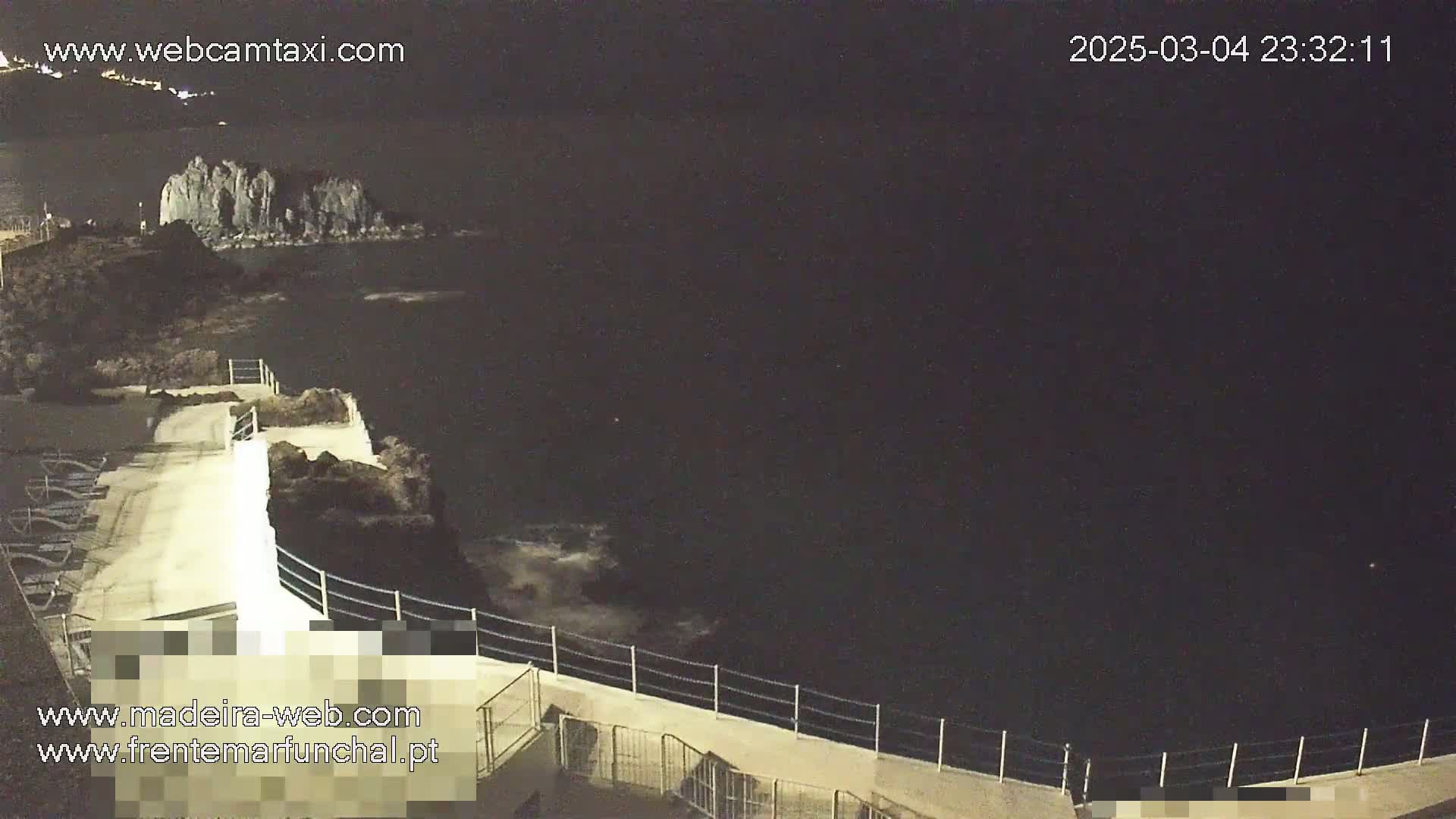 A nighttime view of a rocky coastline with a walkway and railing overlooking dark water with some small waves.