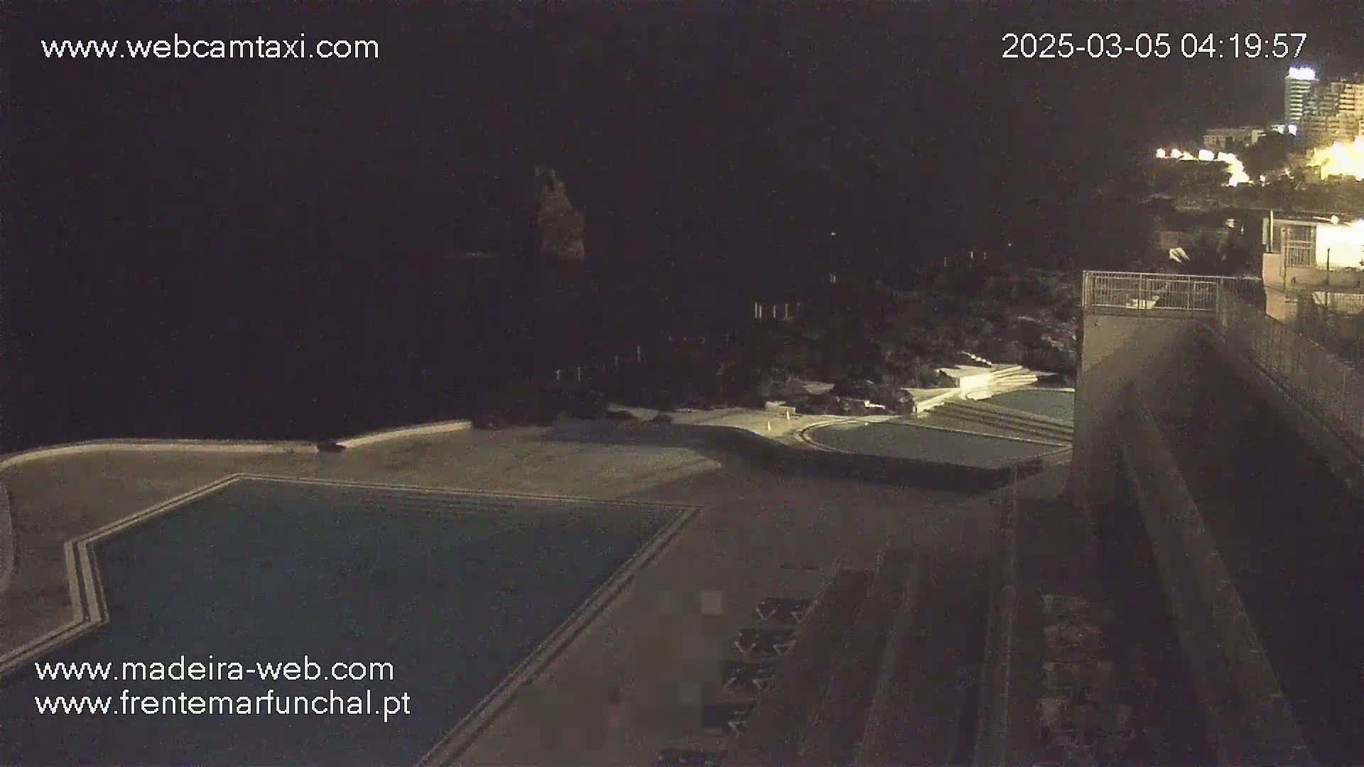 A nighttime aerial view shows an empty swimming pool and surrounding patio furniture next to a dark building and distant city lights.