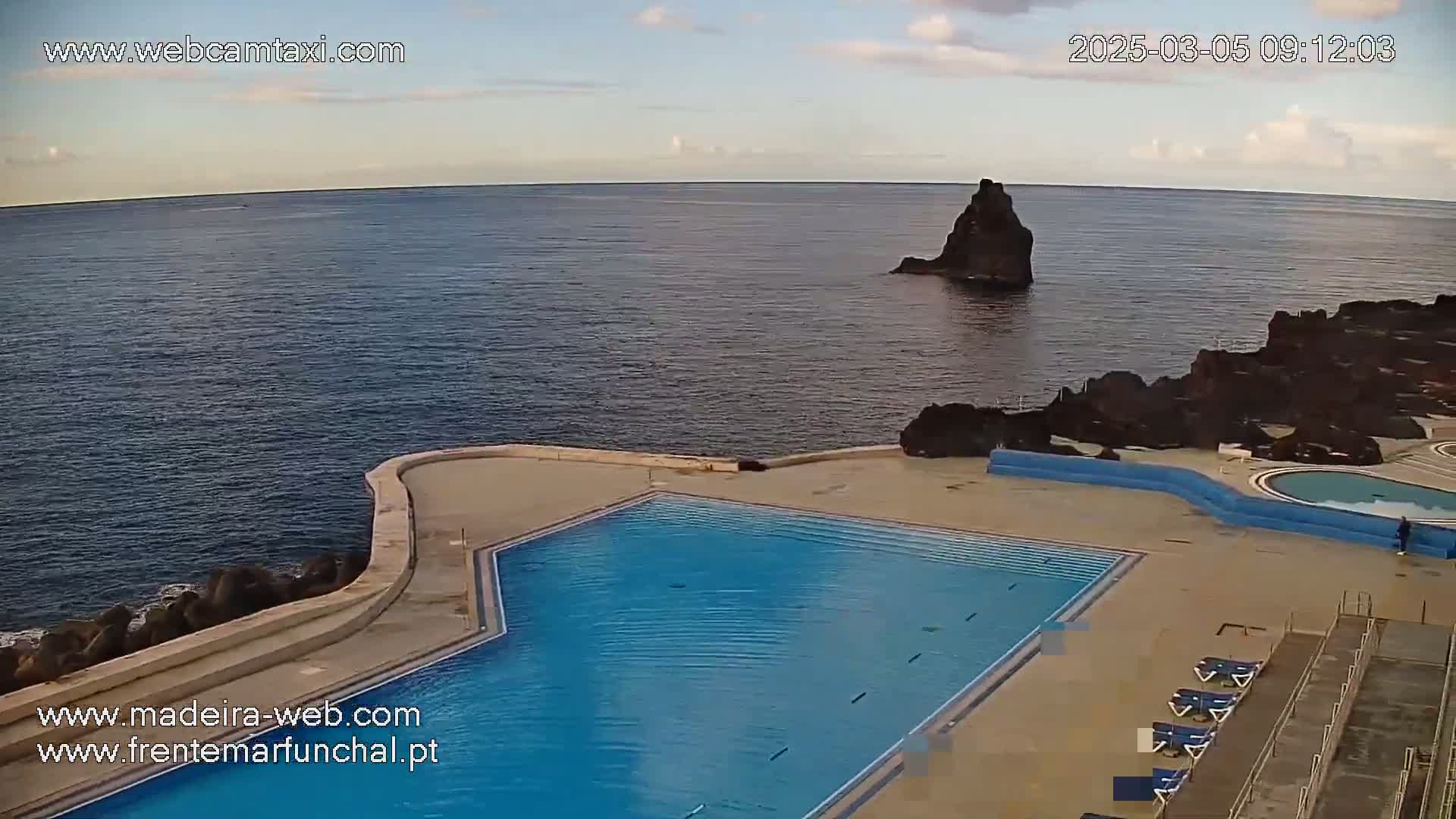 An outdoor pool complex sits on a rocky coast overlooking a calm ocean under a partly cloudy sky.