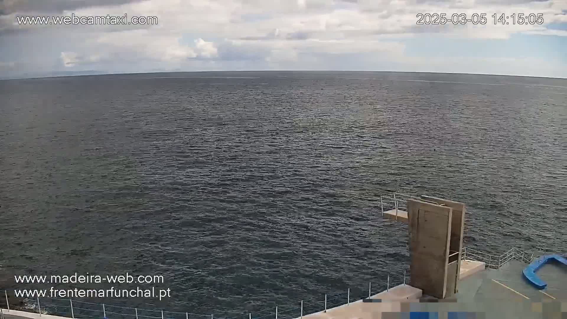 A mostly cloudy sky overlooks a calm, dark-grey ocean with a diving platform on a concrete area in the foreground.