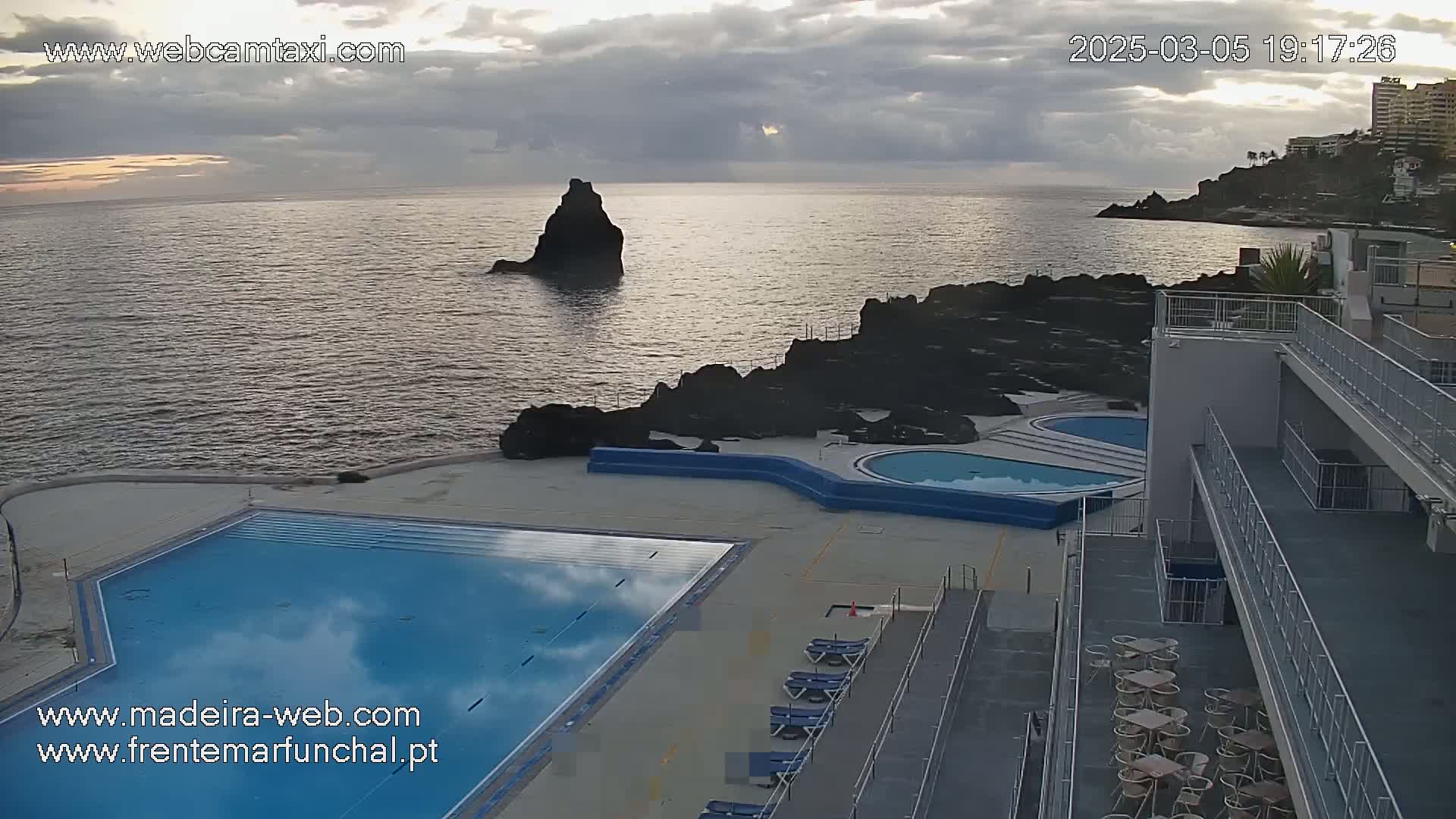 An outdoor pool and patio area overlook a calm ocean with a rocky coastline under a cloudy sky.