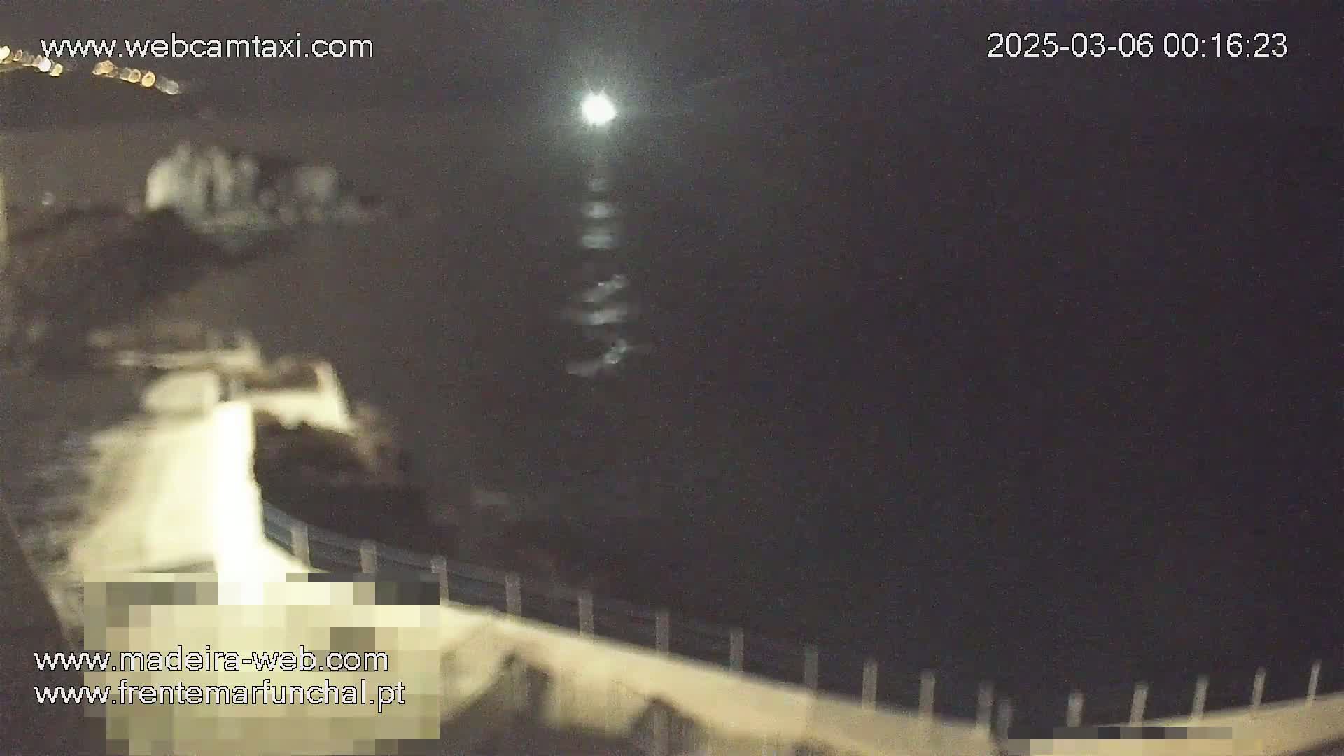 A nighttime view of a dark body of water reflecting a bright light source, with a low wall or barrier in the foreground.