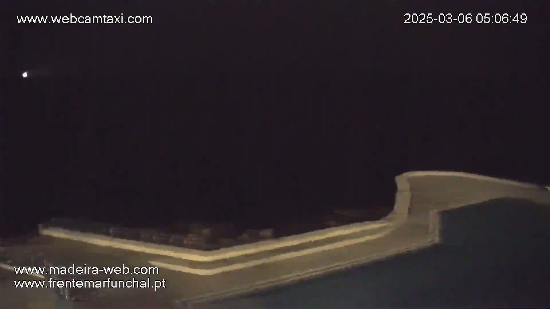 A nighttime view shows a section of a light-colored structure, possibly a wall or railing, against a dark sky with a bright object moving across the upper-left.