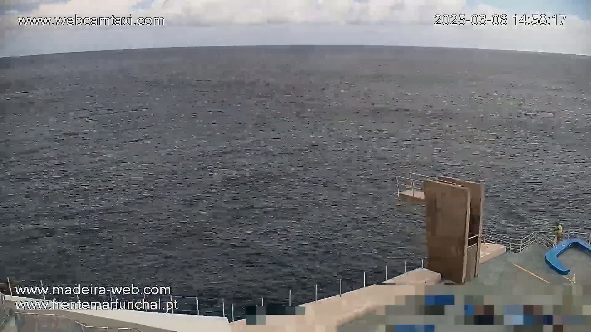 A mostly cloudy day shows a dark body of water with a diving platform and a person standing near a blue structure at the water's edge.